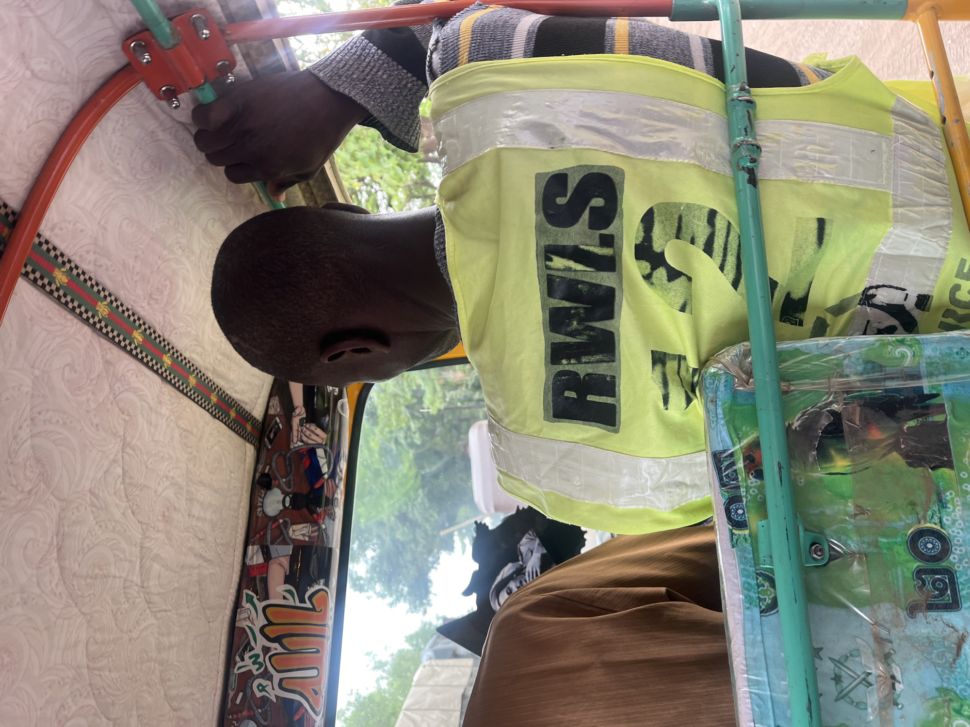 A close-up, rotated portrait-oriented shot shows a person inside what appears to be a small vehicle or rickshaw, likely in Karmajiji, Nigeria. The individual, seen from the back of the head and shoulder, has short, dark hair and dark skin. They are wearing a light green or yellow safety vest with reflective strips, and a striped long-sleeved shirt underneath. The vest clearly displays the black letters "RWIS" and a partially visible word, possibly "FORCE," on the right side.
The person's right hand is visible, gripping a light green metal bar that is part of the vehicle's frame. Another orange-red bar is connected nearby with a metal bracket. The vehicle's interior features a white or off-white quilted fabric lining the ceiling or upper wall. A decorative ribbon with a black and green checkerboard pattern and a red stripe runs along the edge of this lining. Below the ribbon, a dark panel is adorned with numerous colorful stickers and designs.
Through the vehicle's window, a blurry view of green trees and the sky is visible, indicating it is daytime. On the right side of the frame, a light green metal bar stands vertically, and behind it, a patterned item, possibly a mat or cover, is visible, protected by clear plastic.