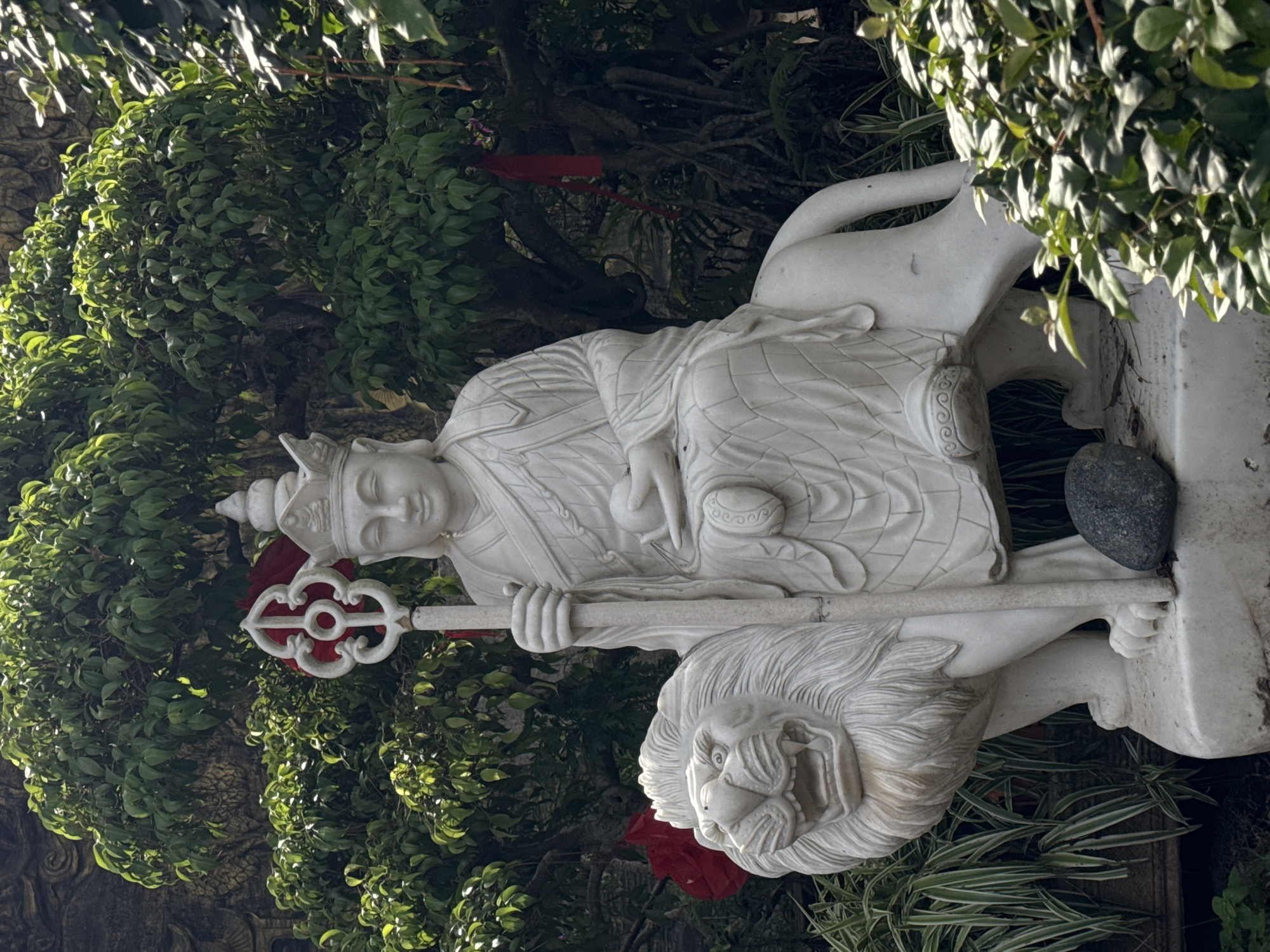 The image is an eye-level, medium shot of a white, marble statue of a seated figure holding a scepter, mounted atop a recumbent lion. The statue is set amidst lush, green foliage and trees, with a dark gray stone resting at the statue's base. The background suggests a garden or park setting. The statue is located in the city of Lâm Viên - Đà Lạt, Vietnam.