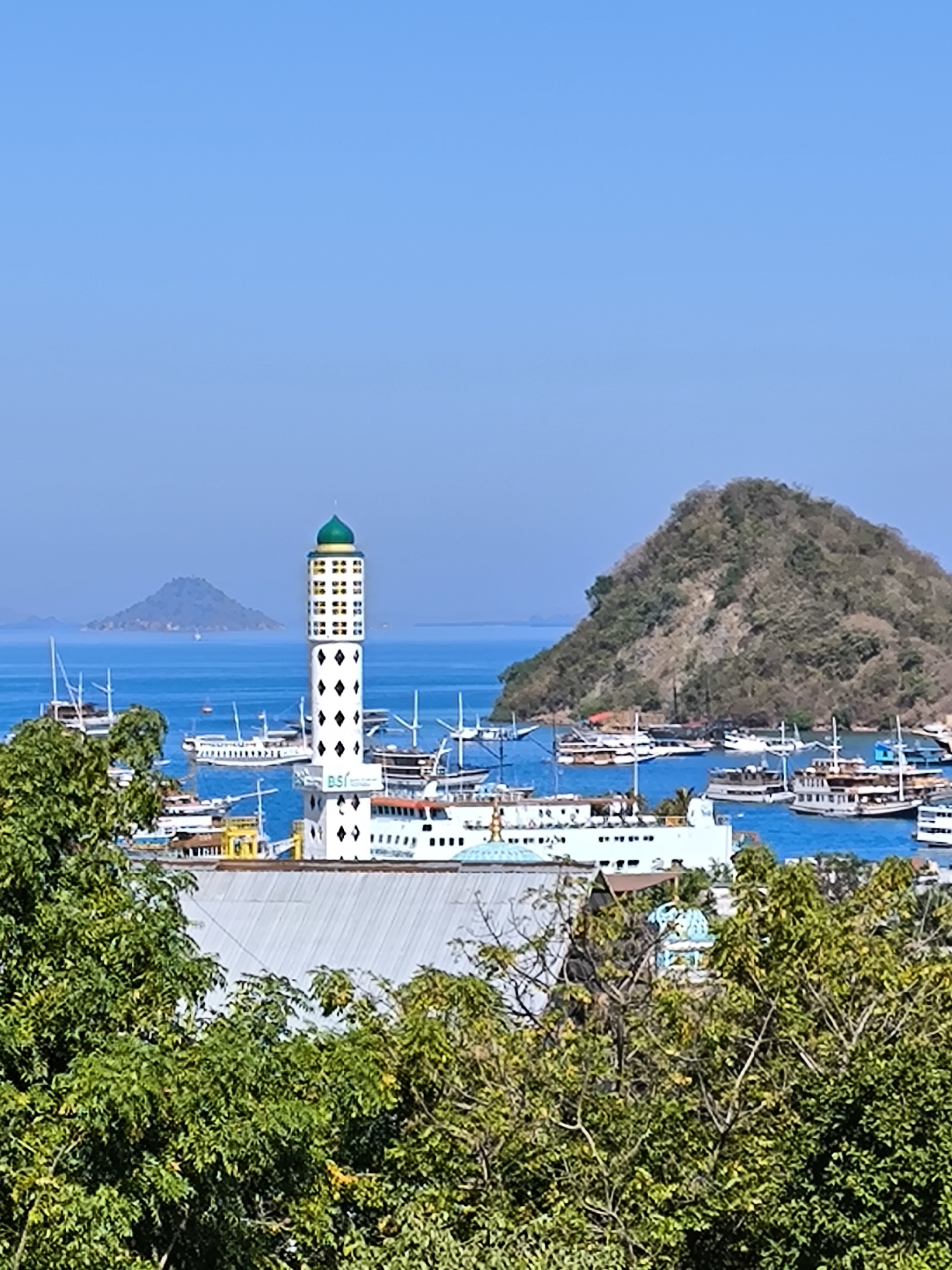 A wide shot captures the harbor of Labuan Bajo, Indonesia, under a clear blue sky. In the foreground, dense green foliage frames the scene. Dominating the midground is a distinctive white tower with black diamond patterns and a green dome, resembling a lighthouse or minaret, with the "BSI" logo visible on its base. Adjacent to it, a white ship with red accents is docked. The harbor is filled with numerous white boats and ships of varying sizes, some with tall masts. In the background, a large, green, tree-covered hill rises from the water, and a smaller, hazy island can be seen in the distance. The water is a vibrant blue, reflecting the clear sky.