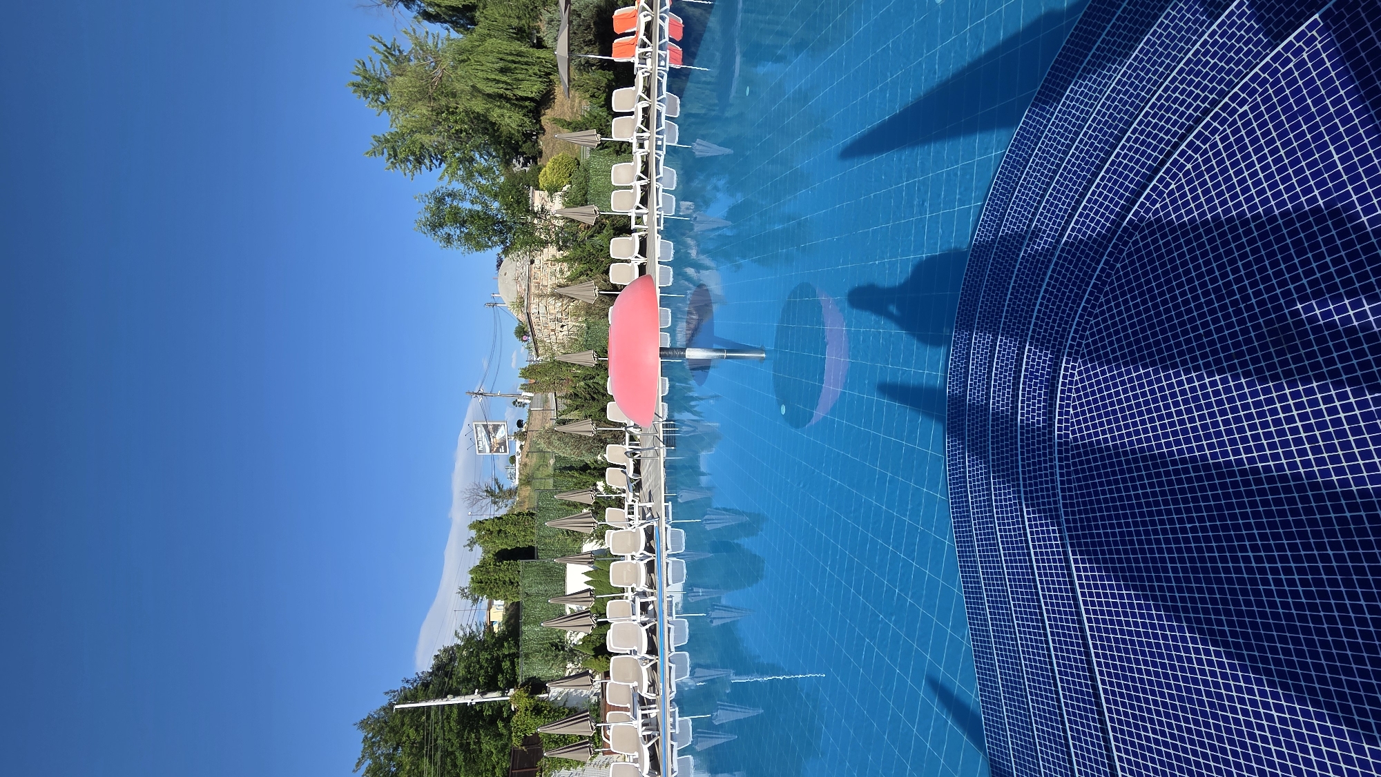 The image shows a swimming pool area at the Aqua Theme park in Bania, Bulgaria. The pool is large and filled with blue water, with a tiled step entrance in the foreground. Surrounding the pool are rows of empty lounge chairs and umbrellas, indicating a scene before or after peak hours. In the center of the pool, a pink mushroom-shaped umbrella provides shade. The background features lush green trees, a stone building with a dome, and a billboard advertising the park. The sky is a clear, bright blue, suggesting a sunny day. Shadows of people and the umbrella are visible on the pool floor, hinting at the presence of visitors.
