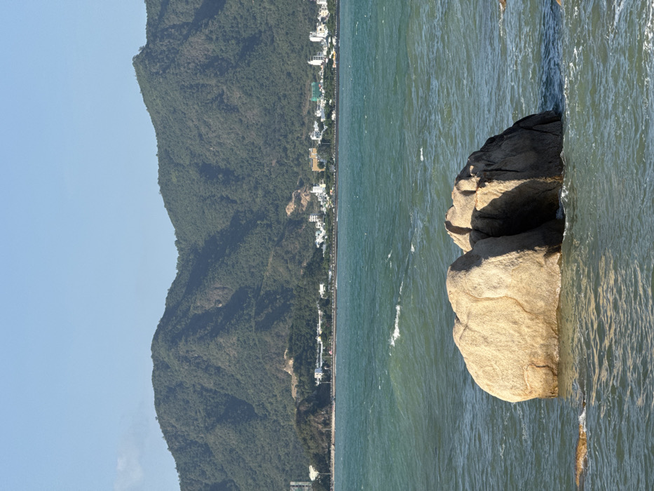 The image displays a coastal scene in Nha Trang, Vietnam. In the immediate foreground, several large, light-colored granite boulders are partially submerged in agitated, teal-green ocean water, with visible waves and white foam. The midground features the expansive ocean. Along the distant coastline, a segment of the city is visible, characterized by a line of white and light-colored buildings situated behind a dark-lined promenade or road. The background is dominated by a range of large, densely vegetated green mountains and hills, silhouetted against a clear, light blue sky. No individuals or specific actions are discernible.