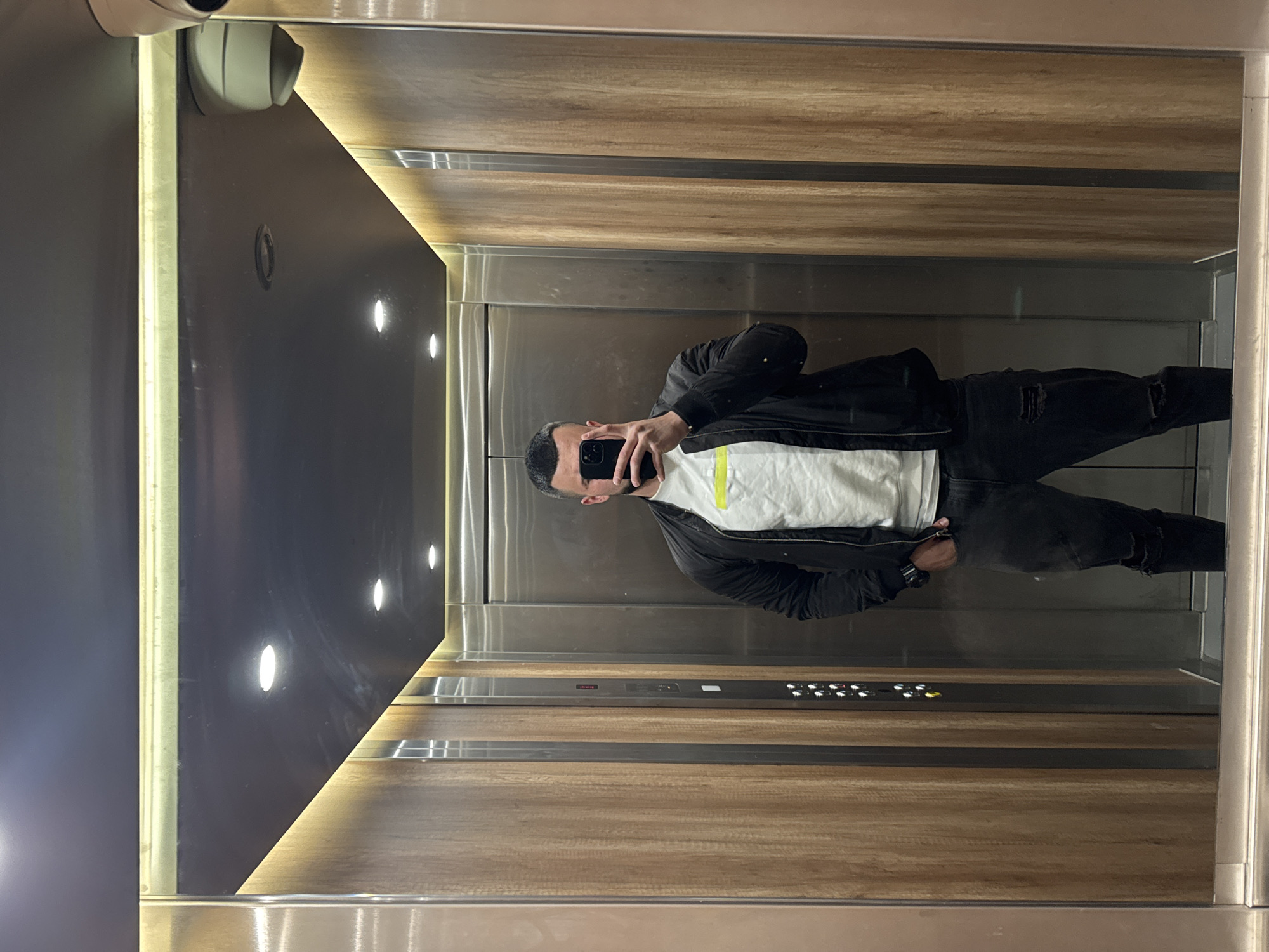 A man is taking a photo of himself in an elevator. The elevator has dark, reflective panels and wood trim. There is a control panel with buttons. The man is wearing a white t-shirt, black jacket, and black jeans. The location is likely within Sofia, Bulgaria, based on the query.
