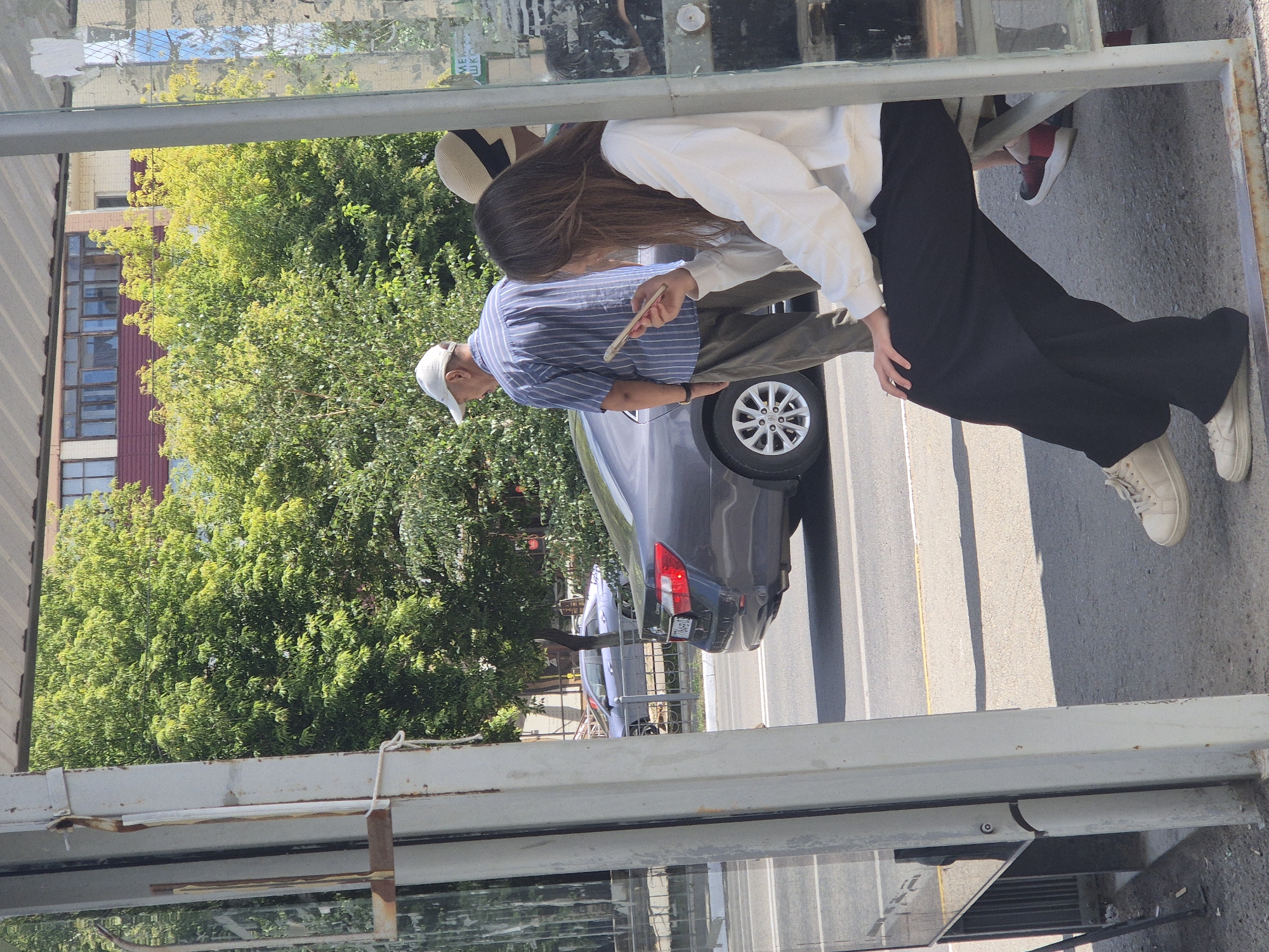 A young woman with long brown hair, wearing a white sweatshirt and black pants, sits on a bench at a bus stop, looking at her phone. She is wearing white sneakers. To her left, an older man in a striped blue and white shirt and khaki pants stands looking away from the camera. Behind him, a dark grey car is parked on the street. A light blue car is visible further down the street. The bus stop has a metal frame and glass panels. Trees and a building with a red roof are in the background. The scene appears to be on a sunny day.