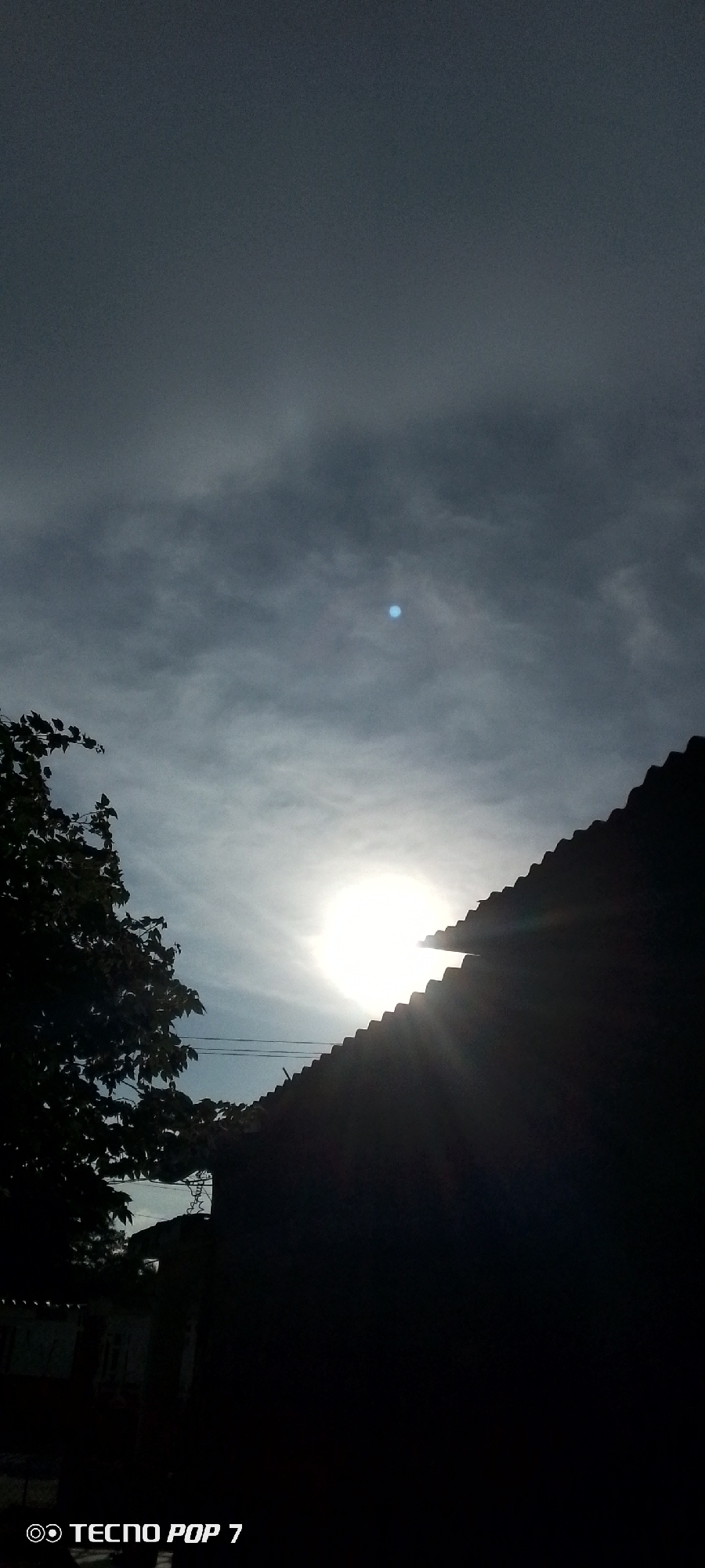 A low-angle, vertical outdoor shot captures a bright sun partially obscured by the jagged edge of a dark, corrugated roof. Sunlight beams outward from behind the roofline, creating visible rays. To the left, the silhouette of a leafy tree frames the scene. The sky is overcast with thin, wispy clouds, and a small, faint bluish-white disc is visible in the upper portion of the sky. The lower part of the image is very dark, showing a sliver of what appears to be buildings and the text "TECNO POP 7" in white at the bottom left corner. The scene is likely taken in Altagracia, Venezuela, given the watermark.