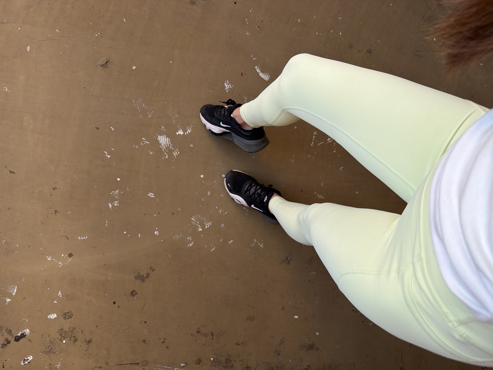An overhead shot depicts a person wearing light yellow leggings and black athletic shoes with white accents, sitting on a brown concrete surface that shows signs of wear and tear with scattered white marks. The person wears a white shirt. The location is assumed to be in the city of Sofia, Bulgaria.