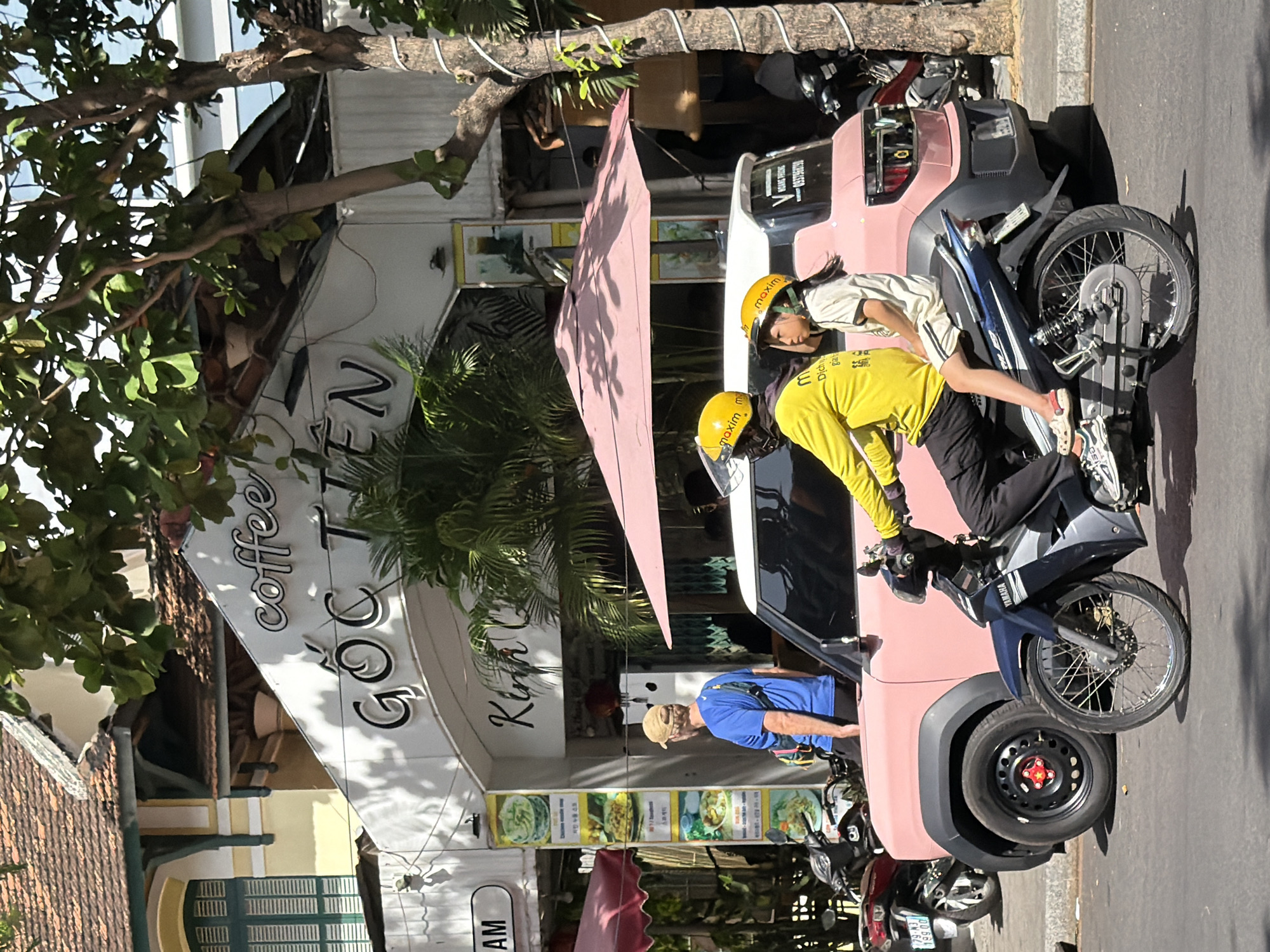 A man and a young girl, both wearing yellow helmets, are riding a motorcycle on a street in Nha Trang, Vietnam. The man is driving and wearing a yellow long-sleeved shirt and black pants, while the girl is a passenger wearing a white t-shirt and shorts. They are positioned in front of a pink car. In the background, there is a building with a sign that reads "GOC TIEN Coffee". The building has a tiled roof and green shutters on one window. There are also trees with green leaves casting shadows on the ground. Several other motorcycles are parked on the side of the street. The scene appears to be in a commercial or urban area.