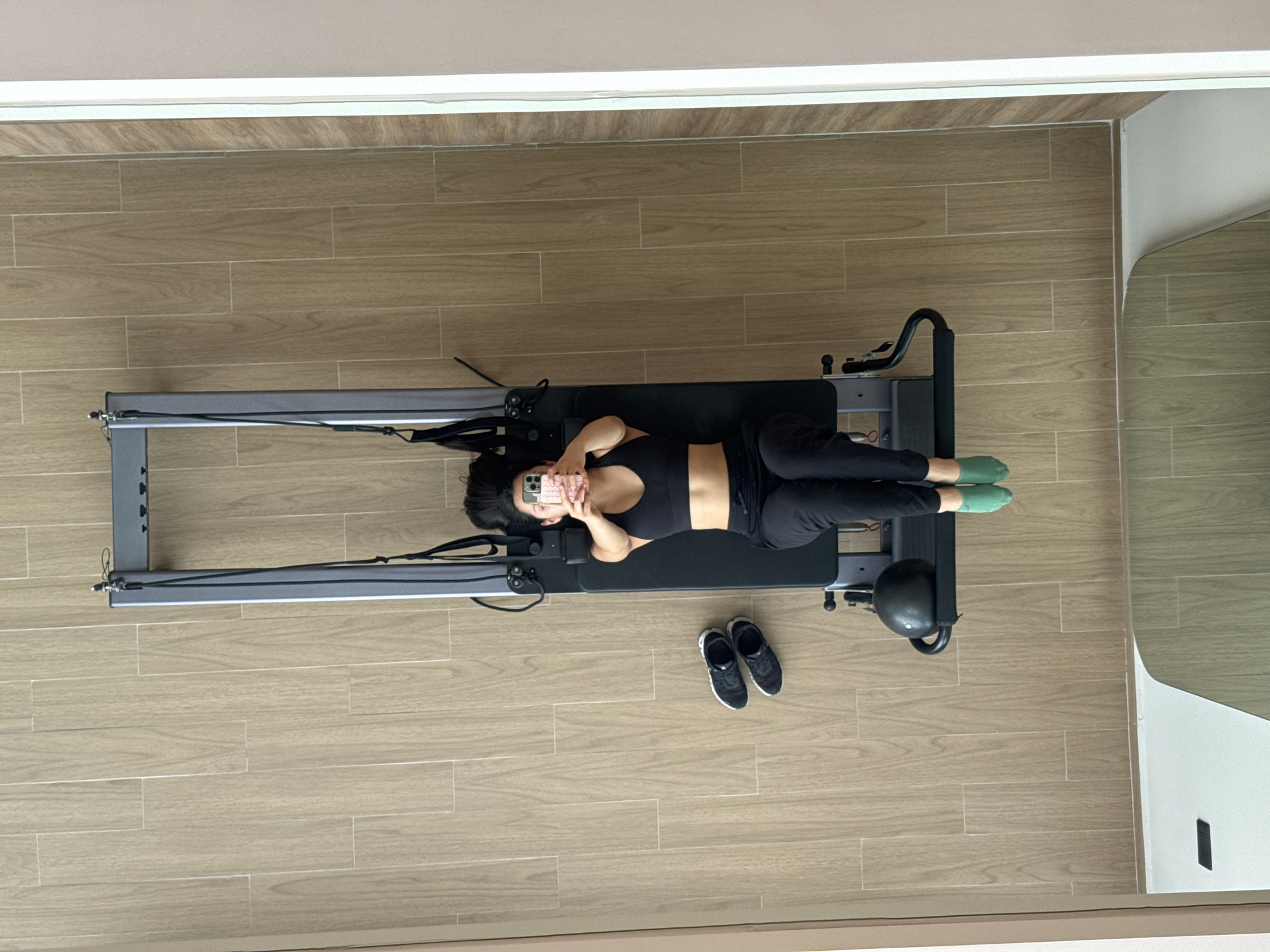 A top-down shot shows a woman lying on a pilates reformer in what appears to be a residential setting with light brown wood-look tile flooring. The woman is wearing a black sports bra and black sweatpants, with green socks. She is holding a mobile phone in front of her face, presumably taking a picture. To the right of the reformer, near her feet, are a pair of black and white athletic shoes. The room has neutral-colored walls, and the lighting is bright. The location is indicated as San Bernardino Tlaxcalancingo, Mexico.