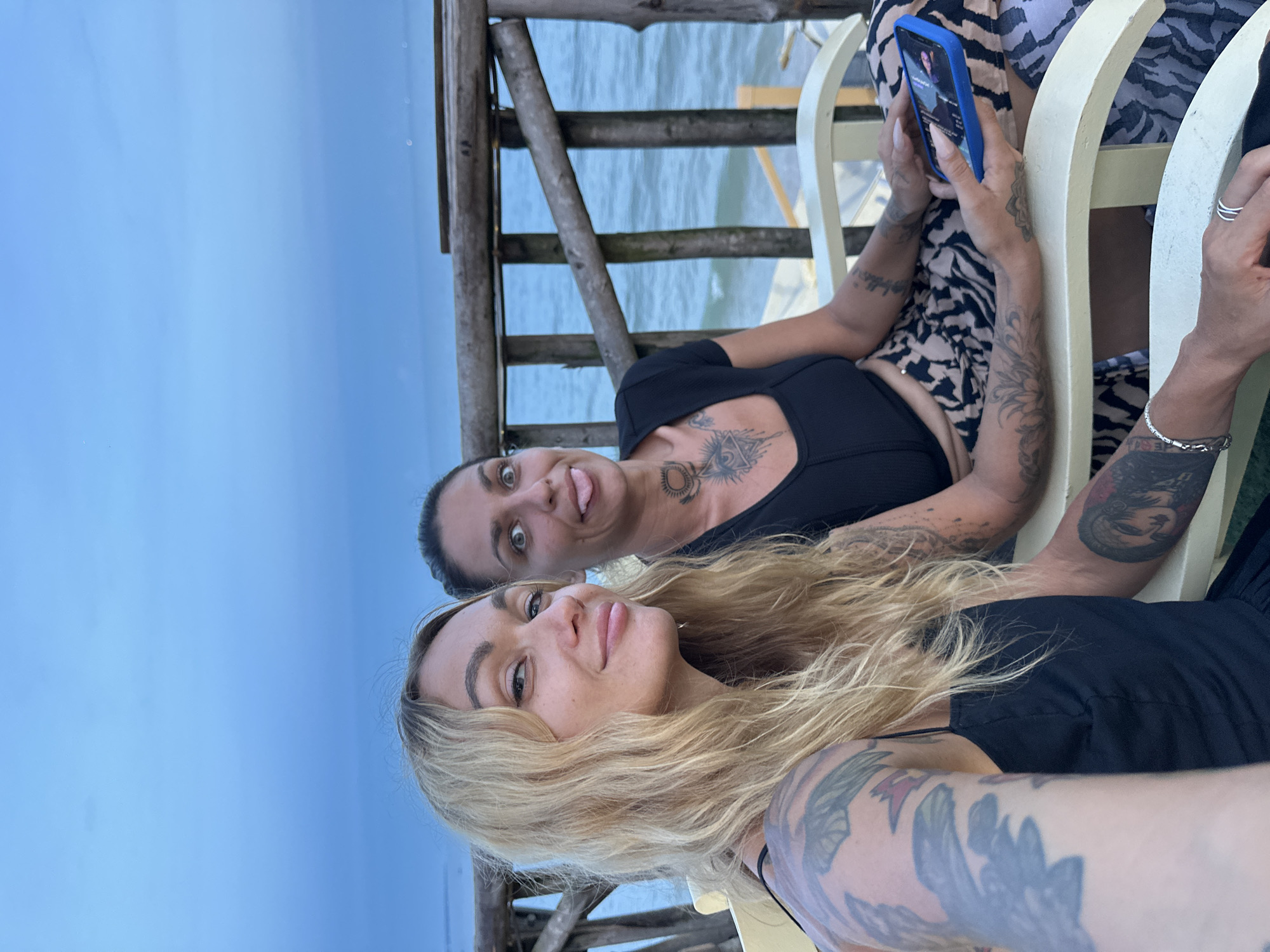 In this vertical, low-angle shot, two women are photographed in an outdoor setting by the water, possibly during the day or early evening, with the sky a pale blue. They are sitting on a light-colored bench on a wooden structure overlooking the water. The woman in the foreground has blonde, wavy hair and is wearing a black top, with a large tattoo on her arm. She is smiling at the camera. The woman behind her, with darker hair, is also looking at the camera with a playful expression, sticking her tongue out. She is wearing a black top with a tattoo visible on her chest. She is holding a blue smartphone in her right hand, and her left arm is resting on the back of the bench. The background shows the calm water and a wooden railing. The overall mood is casual and relaxed.