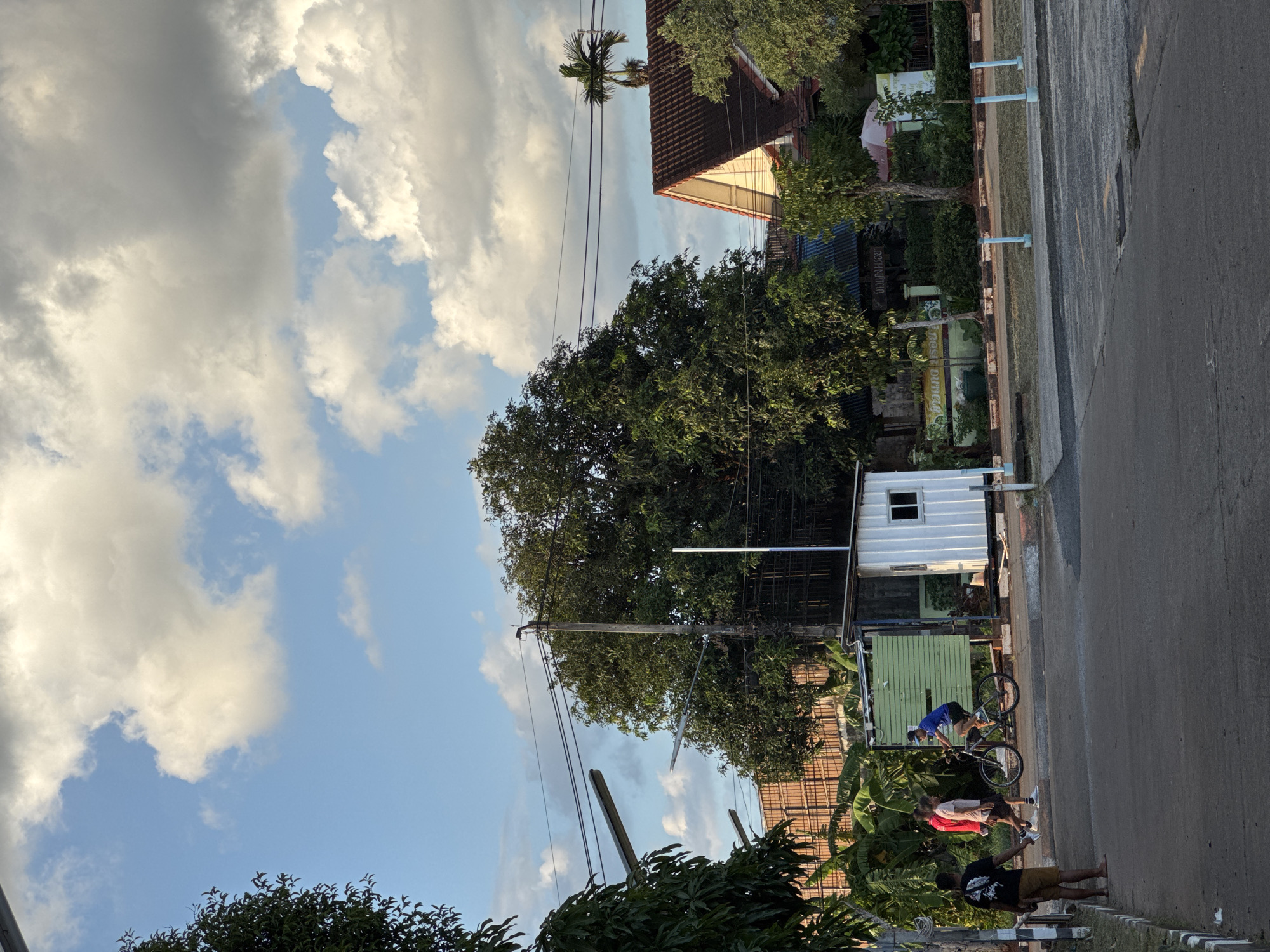 In the city of Surat Thani City Municipality, Thailand, a paved road intersects with a sidewalk. The environment is characterized by lush greenery, including trees and banana plants, under a bright blue sky dotted with fluffy white clouds. On the right side of the road, a residential house with a terracotta tiled roof is visible. Adjacent to it, a small white building with a window stands, possibly a guardhouse or a small office.

Several individuals are present in the scene. On the sidewalk to the left of the road, three people are walking, one wearing a black t-shirt and shorts, another in a red shirt and shorts, and the third in a light-colored t-shirt and shorts. A person on a bicycle, wearing a blue shirt and black shorts, is riding on the road. Another individual stands on the left edge of the sidewalk, wearing a black t-shirt and brown shorts.

The scene appears to be a typical street in a moderately developed area, with a mix of residential buildings and natural vegetation. The lighting suggests it is either early morning or late afternoon, with the sun casting long shadows. Streetlights are mounted on poles along the road, indicating the presence of infrastructure for illumination. The overall impression is one of a serene, everyday moment captured in a tropical setting.