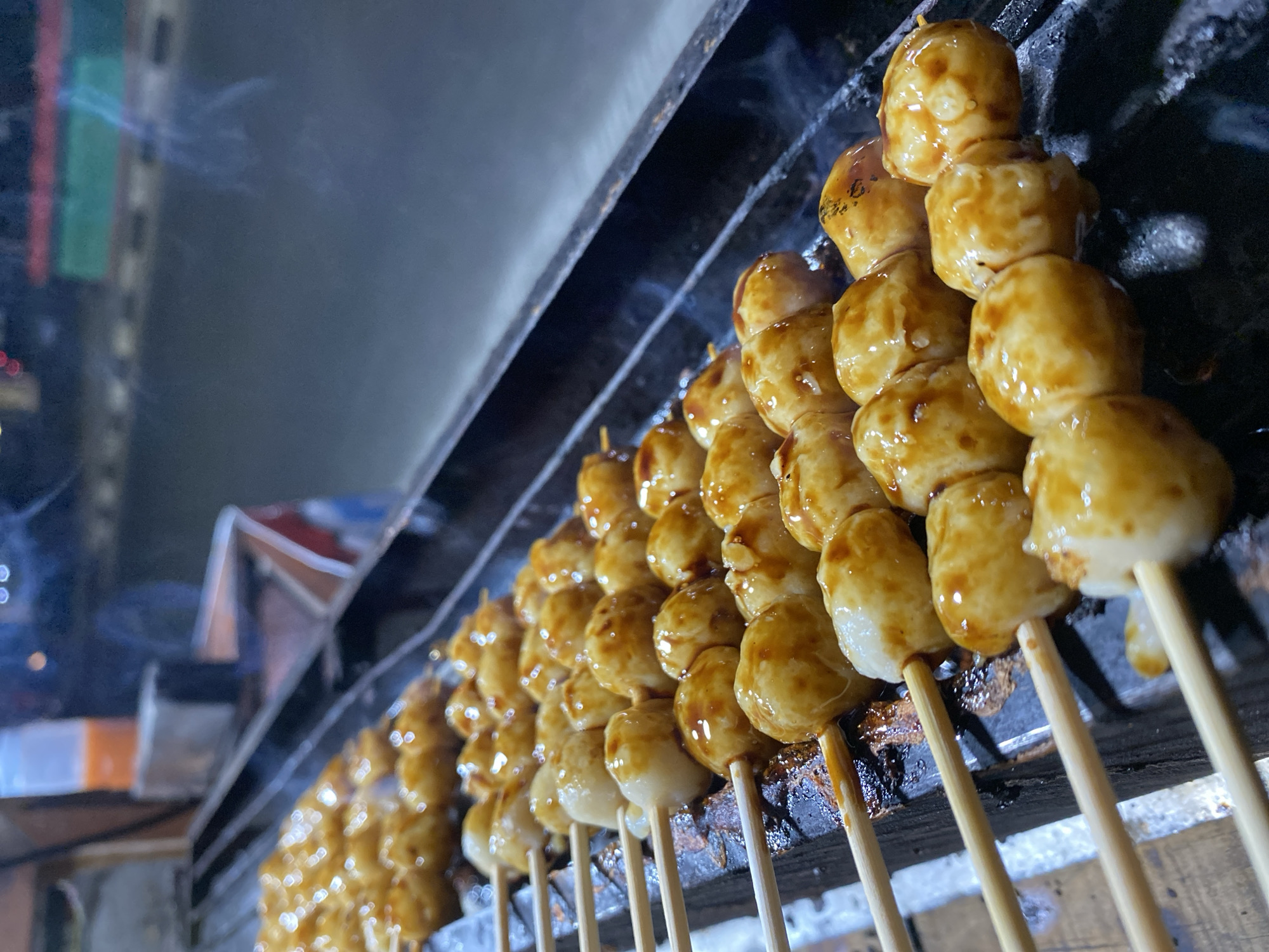 This is a close-up, eye-level outdoor shot of several skewers of golden-brown meatballs being grilled at night, likely on a street food stall in Kasihan, Indonesia. The meatballs are glistening with a dark, possibly sweet sauce and some are lightly charred. Wisps of steam rise from the grill. In the background, out of focus, are blurry lights and what appears to be a road with passing vehicles, suggesting an urban or suburban setting. The grill is made of dark metal with visible grates, and there is some charring on the surface. Wooden skewers hold the meatballs in rows.