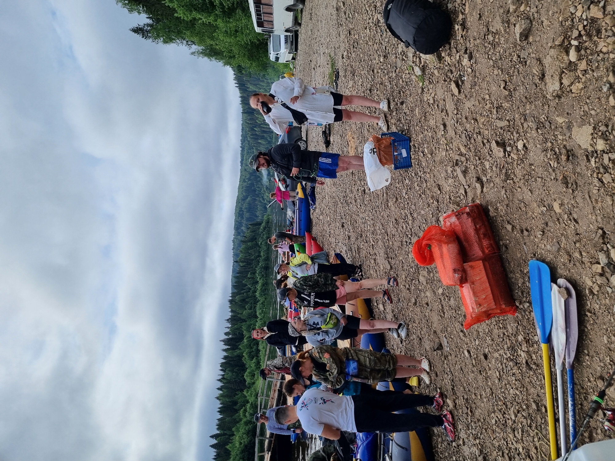 This image depicts a group of people gathered on a rocky shore next to a river in Visherogorsk, Russia. The sky is overcast with scattered clouds. In the background, a dense forest lines the opposite bank of the river. Several inflatable rafts are pulled up onto the shore, suggesting preparations for or the conclusion of a water-based activity, likely rafting. People of various ages are dressed in casual outdoor clothing, some in athletic wear, others in camouflage attire. Some individuals are standing in conversation, while others are observing their surroundings. Objects scattered on the ground include bags, a blue crate, and several paddles lying near the water's edge. In the distance, a white bus and a smaller white van are parked, hinting at transportation support for the group. Two large, orange, netted bundles are stacked on the shore in the foreground, possibly containing gear or supplies.