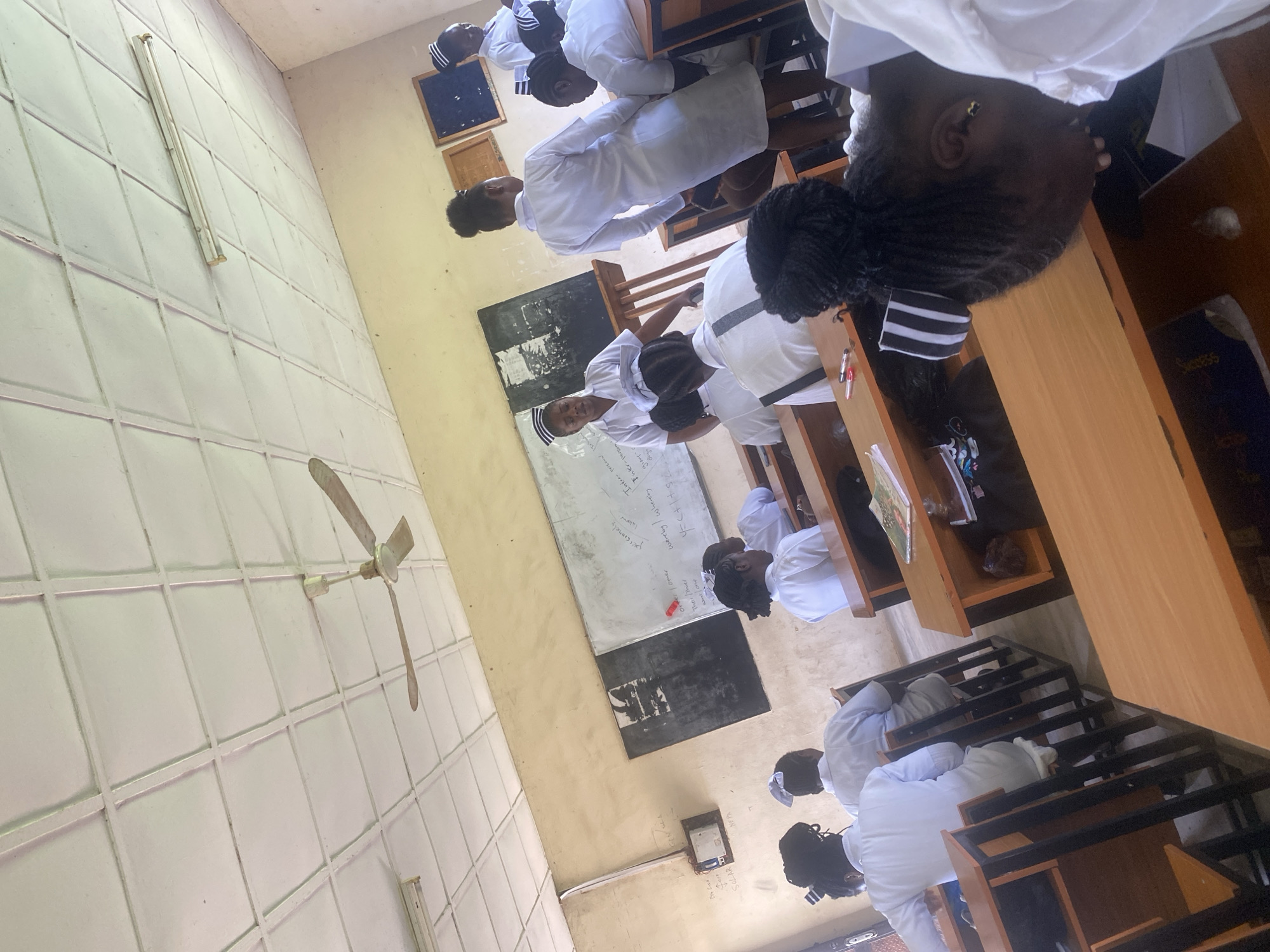 In a classroom setting, several female nursing students in white uniforms and caps are seated at wooden desks. Some students are facing the front, possibly towards a whiteboard visible in the background with writing on it. A female instructor, also in a uniform and cap, stands near the whiteboard, appearing to address the students. The room has a tiled ceiling, a ceiling fan, and fluorescent lighting. Wooden cabinets or notice boards are mounted on the wall. Desks are arranged in rows, and some students have books or notebooks on their desks. The overall environment suggests an educational or training session within a school or hospital facility in Dutsen Kato, Nigeria.