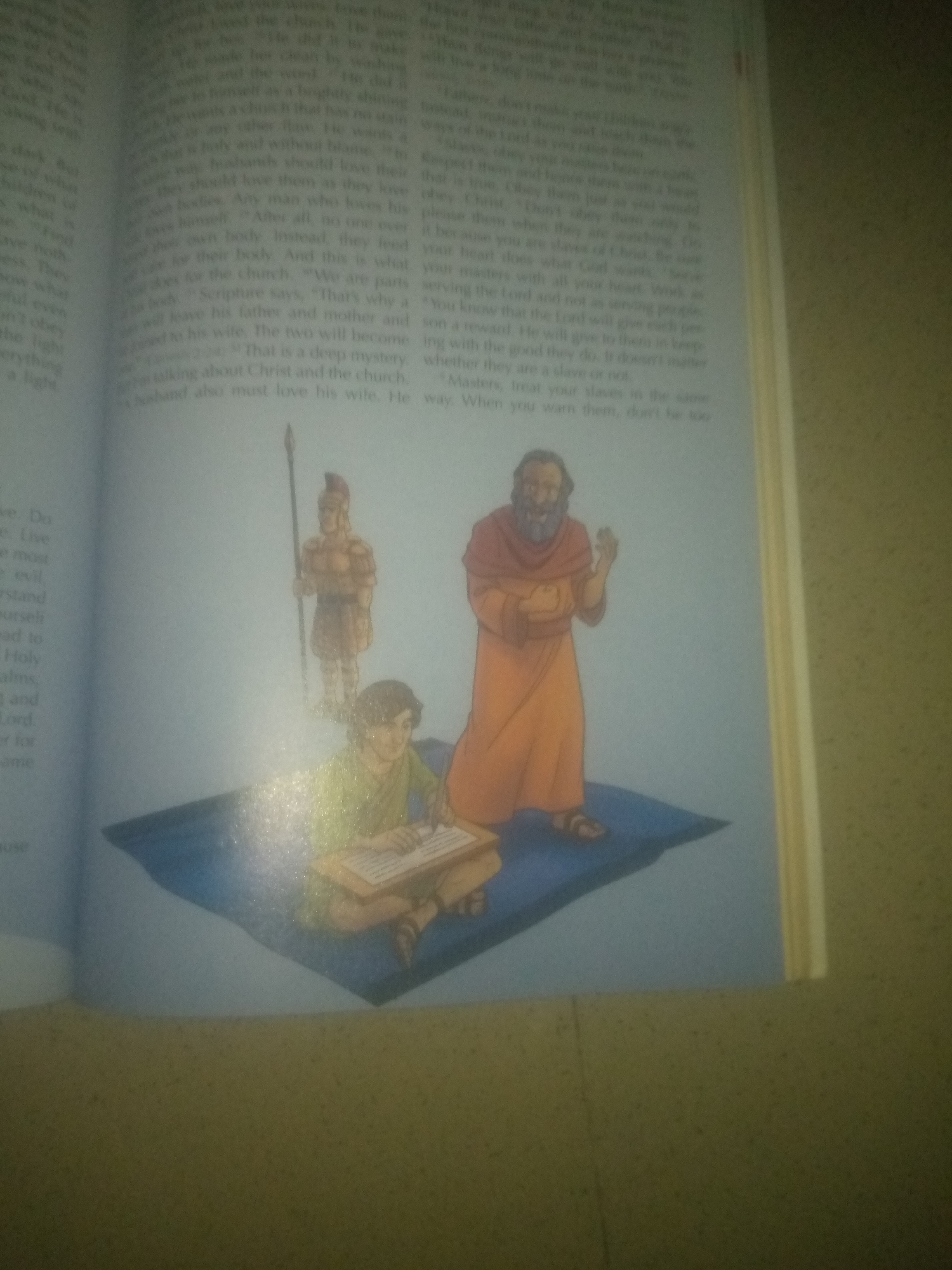 The image is a photograph of an open book with an illustration. The illustration depicts three figures. In the foreground, a young person with dark hair, wearing a green tunic, is seated on a blue rug, writing on a tablet with a stylus. To the right of the young person stands an older person with a beard, wearing an orange robe and sandals, holding a loaf of bread. Behind them, a figure dressed as a Roman soldier stands at attention, holding a spear. The text in the book is partially visible, with the illustration dominating the right side of the frame. The environment appears to be an indoor setting, possibly a room, with the book resting on a surface. There is no indication of a specific location in the city of Jauro Yinu, Nigeria.