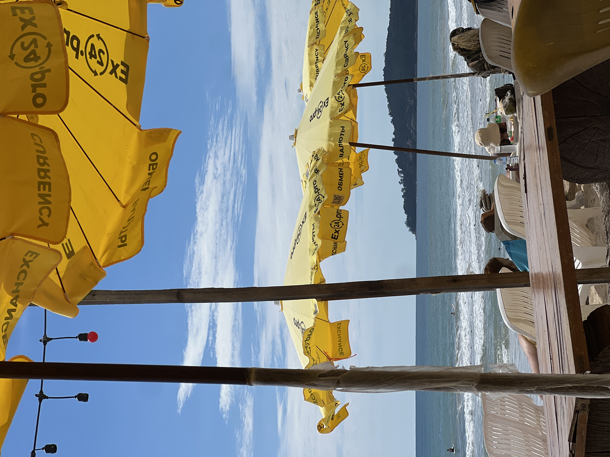 An eye-level, outdoor shot captures a beach scene with yellow umbrellas and a table in the foreground. The umbrellas, adorned with black text that reads "EXCHANGE CURRENCY" and "EXCHANGE PRO," are positioned to provide shade. In the background, the ocean stretches to the horizon, with waves gently rolling onto the shore. A small, tree-covered island is visible in the distance. Several white plastic chairs are scattered around the beach, and a wooden table sits in the immediate foreground, laden with drinks and snacks. A few individuals are visible in the water, and others are seated in the chairs. The sky is a clear blue, dotted with a few white clouds, and the overall atmosphere suggests a relaxed beach day.
