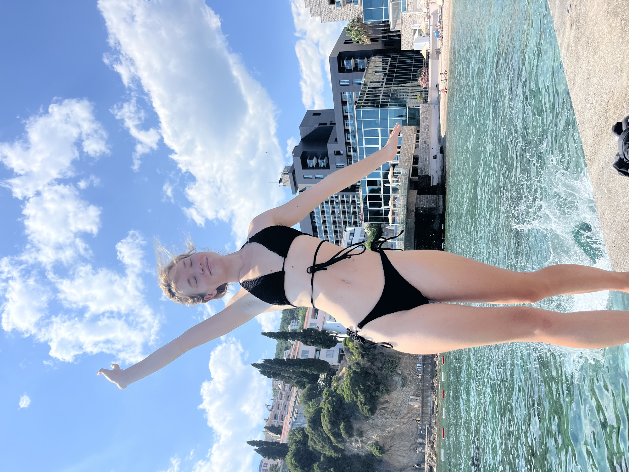 A young woman in a black bikini stands on a concrete pier, her arms outstretched and eyes closed in enjoyment. The sun illuminates her as she faces the camera, her hair windswept. Behind her, the turquoise waters of the Adriatic Sea churn with a splash, indicating a recent jump or movement. The coastline of Budva, Montenegro, unfolds in the background, featuring a lush green hillside dotted with buildings and cypress trees. Modern hotels with dark facades and glass windows rise along the shore, contrasting with the natural landscape. A sandy beach with scattered sunbathers and umbrellas is visible further down the coast. The sky above is a vibrant blue, adorned with fluffy white clouds.
