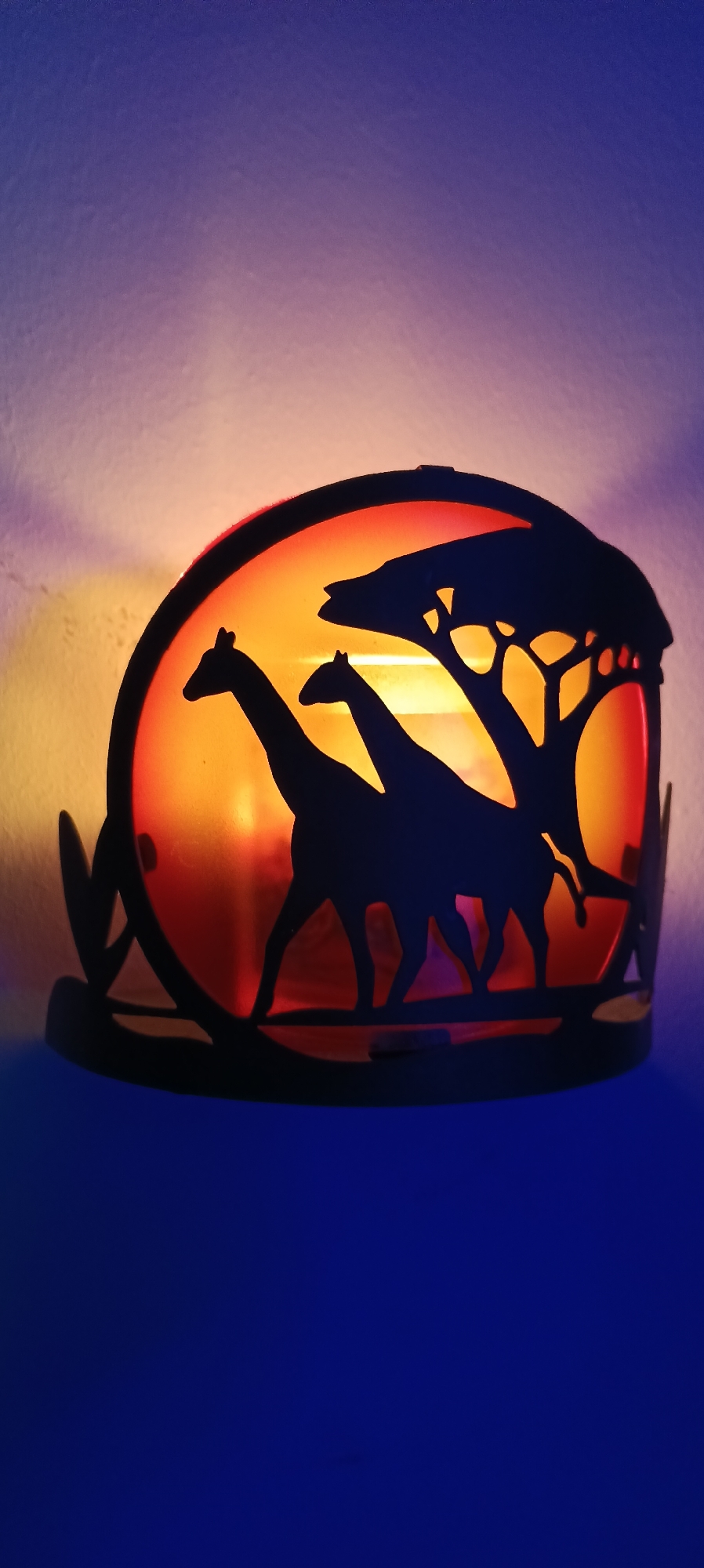 This is a close-up, vertical, eye-level shot of a decorative candle holder shaped like an African savanna scene at dusk. The candle holder is made of black metal and features silhouettes of two giraffes and an acacia tree against a translucent, orange and red background, which is illuminated by a candle within. The background of the image is a gradient of colors, transitioning from a warm yellow-orange at the bottom to a soft purple and blue at the top, likely from ambient lighting. The candle holder itself is placed on a dark blue surface. The image appears to be taken indoors, possibly in a home or a decorative setting. No specific location in Wels, Austria, can be identified from the image.