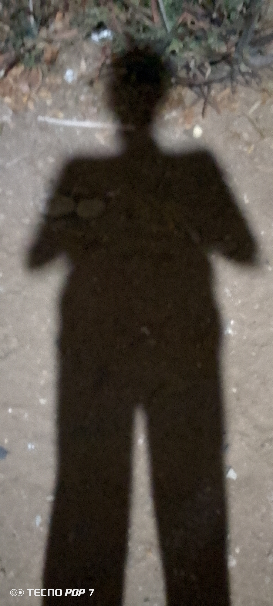 A full-body shadow of a person is projected onto a rough, light-colored surface. The shadow indicates an individual with a distinct head shape, a torso with arms extending slightly outwards, and legs positioned apart. The surface appears to be a ground area composed of small particles. The phrase "TECNO POP 7" is visible in the lower left corner, likely indicating the device used to capture the image. The location of Altagracia, Venezuela is inferred.