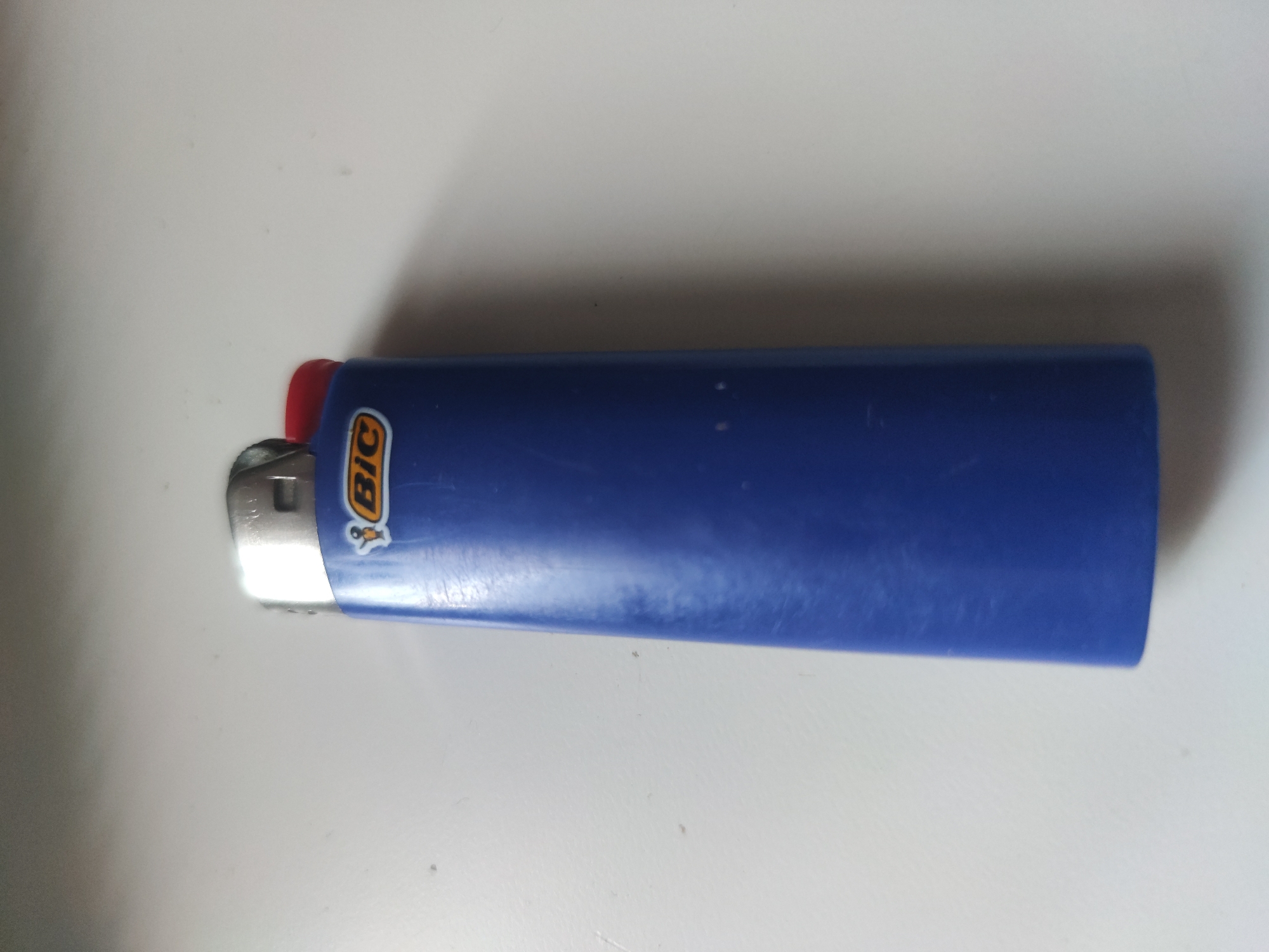 A blue Bic lighter lies on a white surface. The lighter is oriented horizontally with the flint wheel and red button on the left. The Bic logo is visible on the side of the lighter. The location is unknown.