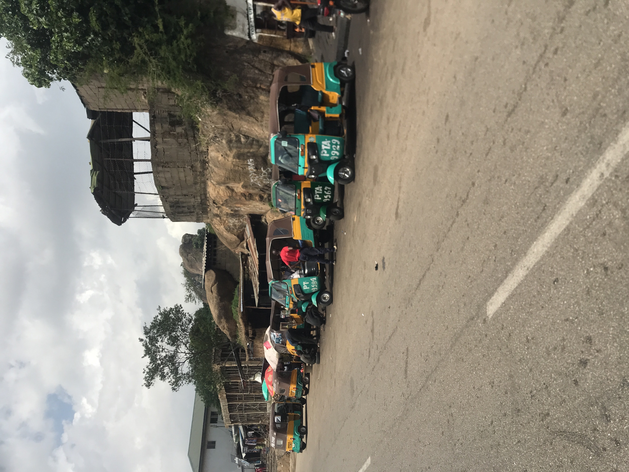 In New Stadium, Nigeria, a lineup of colorful auto-rickshaws, also known as "Keke Napep," are parked along a paved road. The vehicles are predominantly yellow and green, with some featuring brown roofs. Several of these auto-rickshaws have identifying numbers, such as "PTA 9929" and "PTA 9567" painted on their sides.
In the background, a rocky hillside forms the backdrop, with a few trees and structures scattered across it. One structure appears to be a covered pavilion with a weathered roof and wire fencing. To the left, a building with a green roof and white walls is visible, alongside market stalls with various goods displayed.
Several individuals are present. One person in a red shirt stands near the front of a green and yellow auto-rickshaw, while others are seen near the parked vehicles, some possibly attending to the vehicles or engaging in other activities. The sky above is partly cloudy. The scene captures a typical street view of transportation and commerce in this Nigerian city.