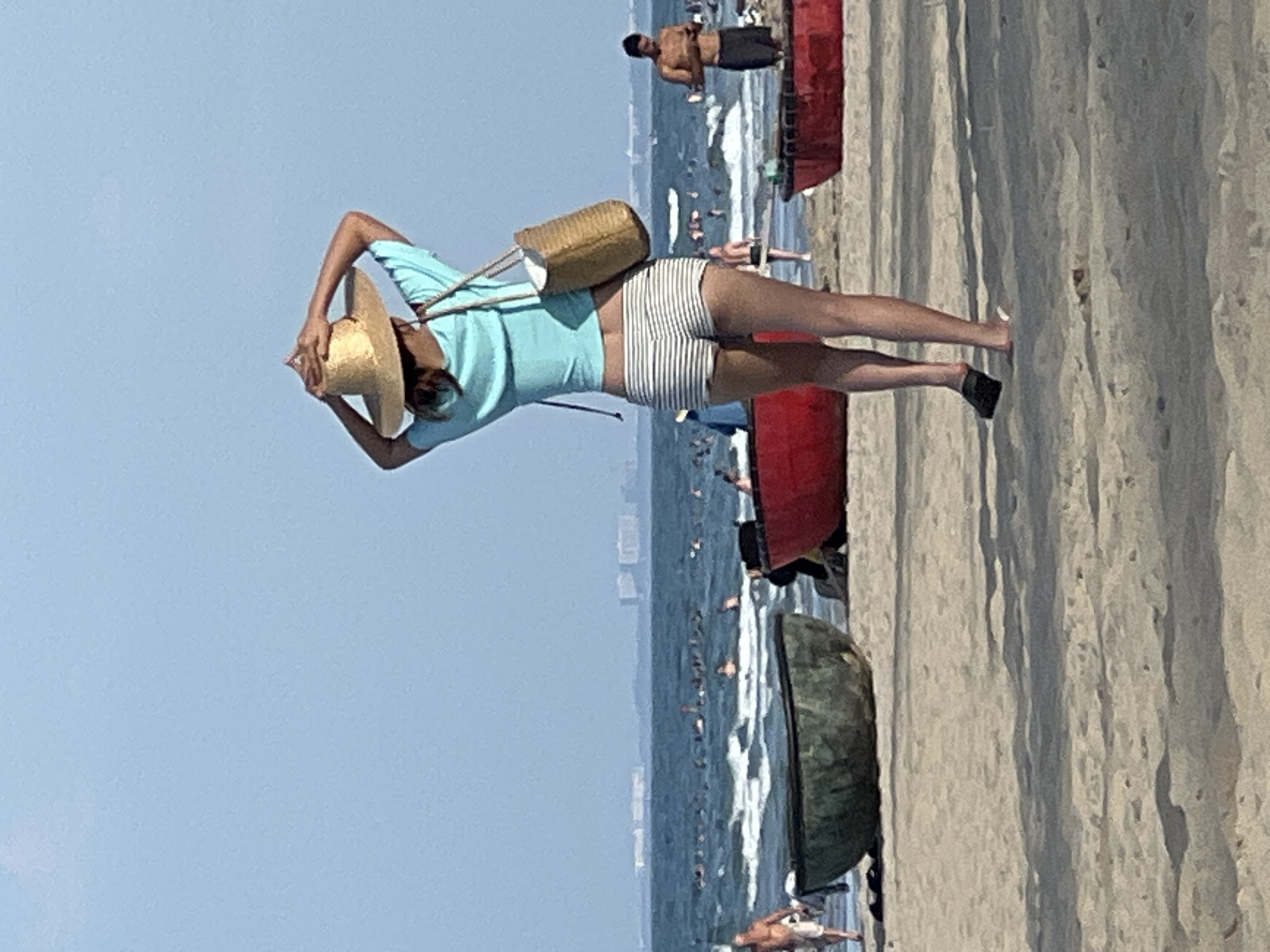 In Đà Nẵng, Vietnam, a woman with brown hair is standing on a sandy beach, facing away from the camera. She is wearing a light blue cropped t-shirt, striped shorts, and a straw hat that she is adjusting with her hands. A woven tote bag hangs from her right shoulder. In the background, there is a calm ocean with small waves and several people swimming. Two round, basket-like boats, one dark green and one red, are visible on the sand in the foreground. The sky is a clear, pale blue. To the right of the woman, a man in black shorts is standing by the water. In the distance, across the ocean, a hazy cityscape with buildings can be seen. The overall scene depicts a relaxed day at the beach.