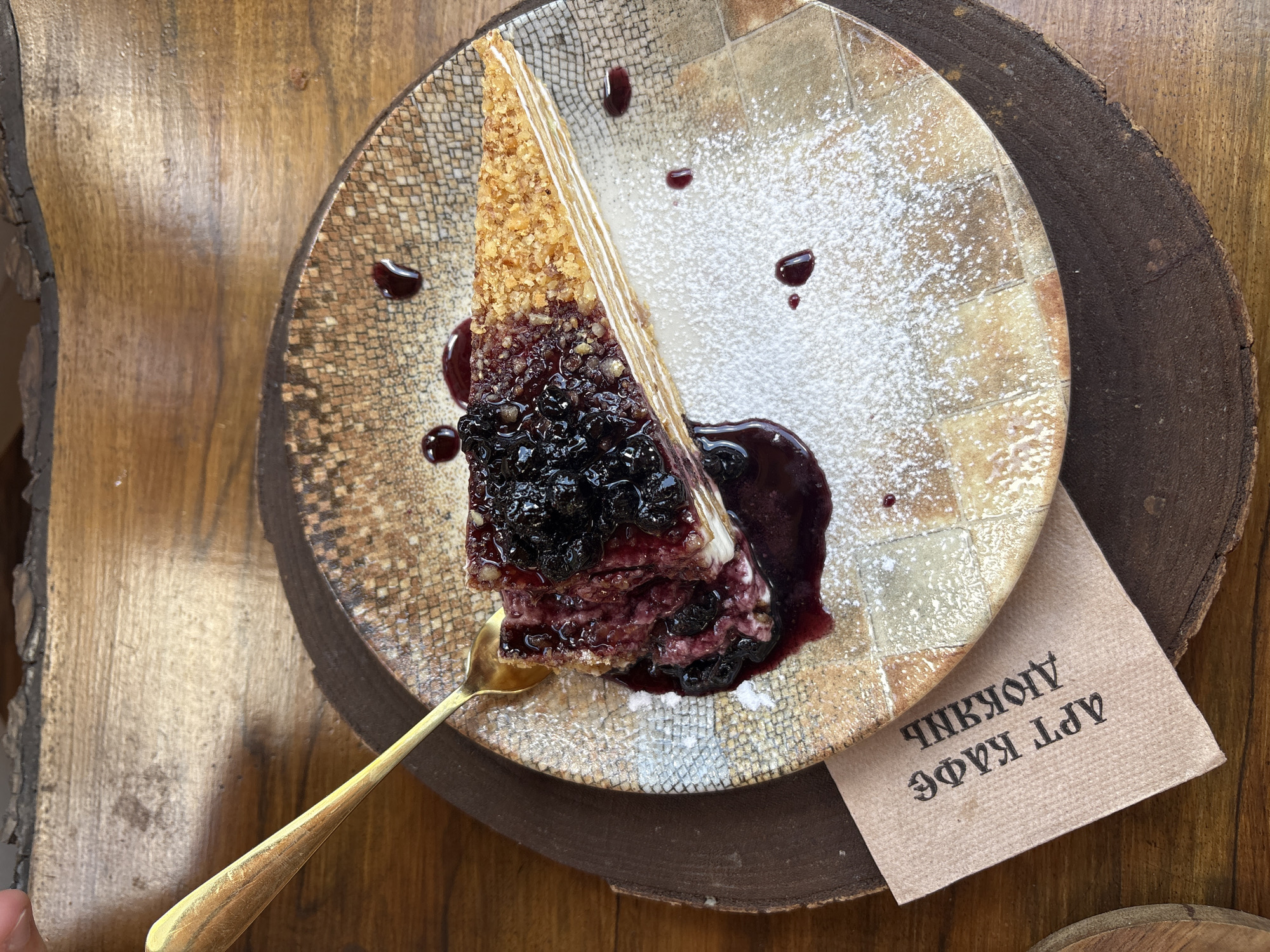 A slice of layered cake, topped with blueberries and a berry sauce, sits on a patterned ceramic plate. The plate is placed on a wooden surface, possibly a table. A gold spoon rests on the plate, partially in the cake. A brown paper napkin with Cyrillic text is tucked under the plate. The scene is indoors, likely a cafe, with natural light illuminating the food. The location is the city of Shiroka Laka, Bulgaria.