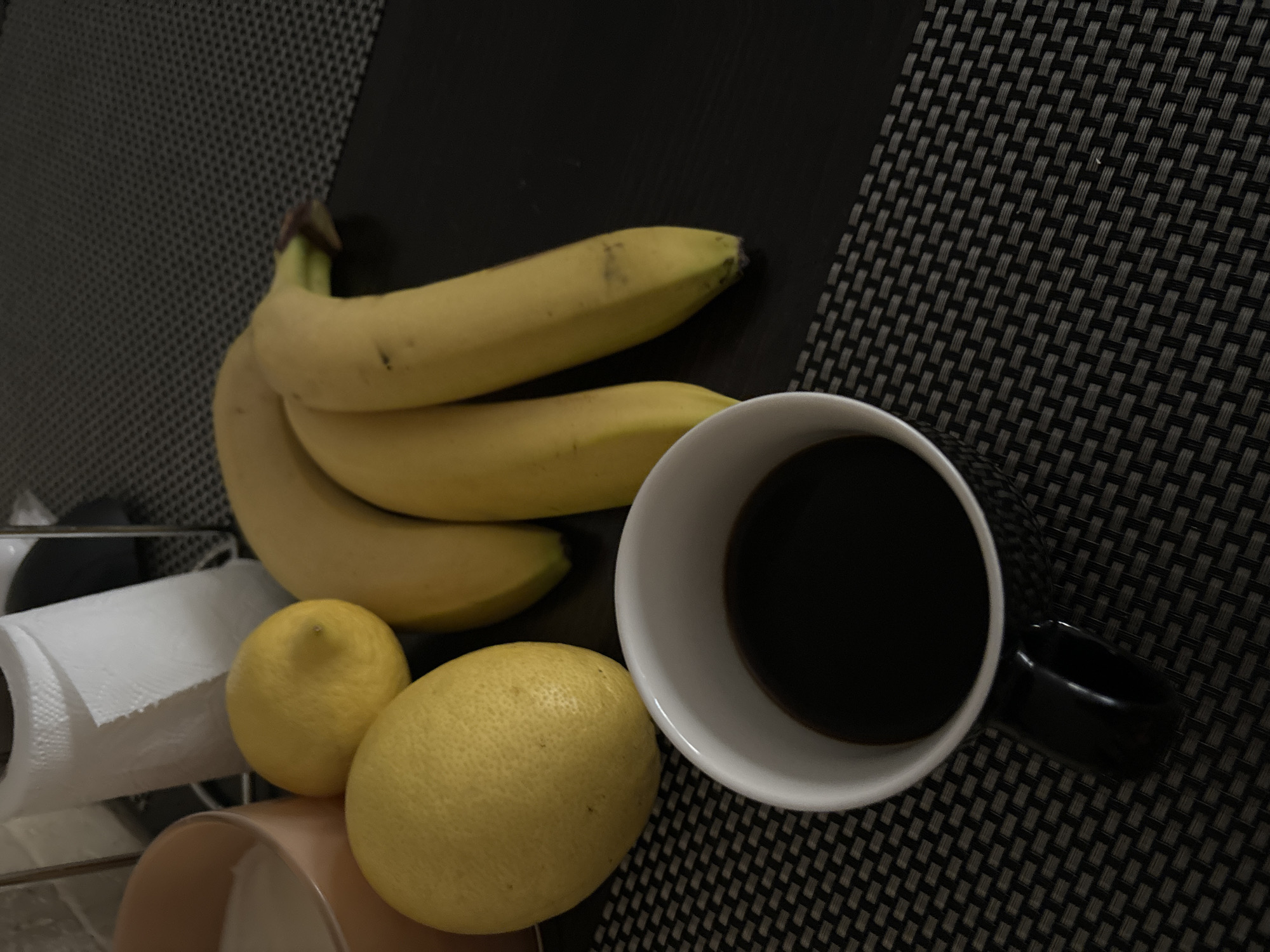 The image shows a still life arrangement on a dark woven surface, likely a table or countertop. There are three bananas, two lemons, and a white mug filled with a dark liquid, probably coffee or tea. A roll of paper towels and a ceramic container are partially visible in the lower left corner. The scene is indoors. The location in Budva, Montenegro cannot be determined from the visual information provided.