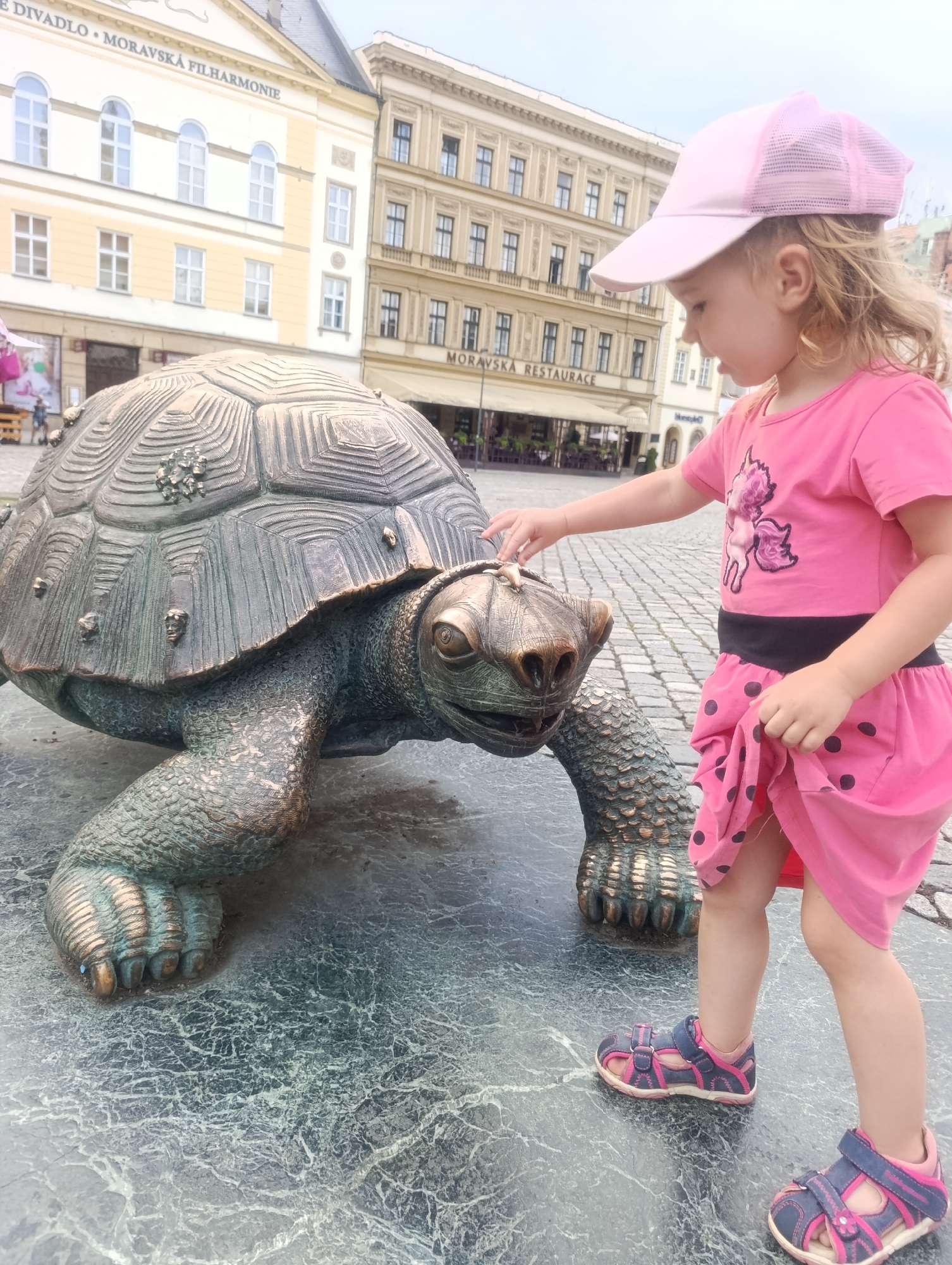 A young girl wearing a pink baseball cap and a pink dress with black polka dots stands on a cobblestone square in Olomouc, Czechia, touching a large bronze turtle sculpture. The turtle sculpture is positioned on a dark, textured stone base. In the background, classical buildings with light-colored facades and arched windows line the square. A sign on one of the buildings reads "MORAVSKÁ FILHARMONIE" and another reads "MORAVSKÁ RESTAURACE". The girl is wearing pink and blue sandals. The sky is overcast.