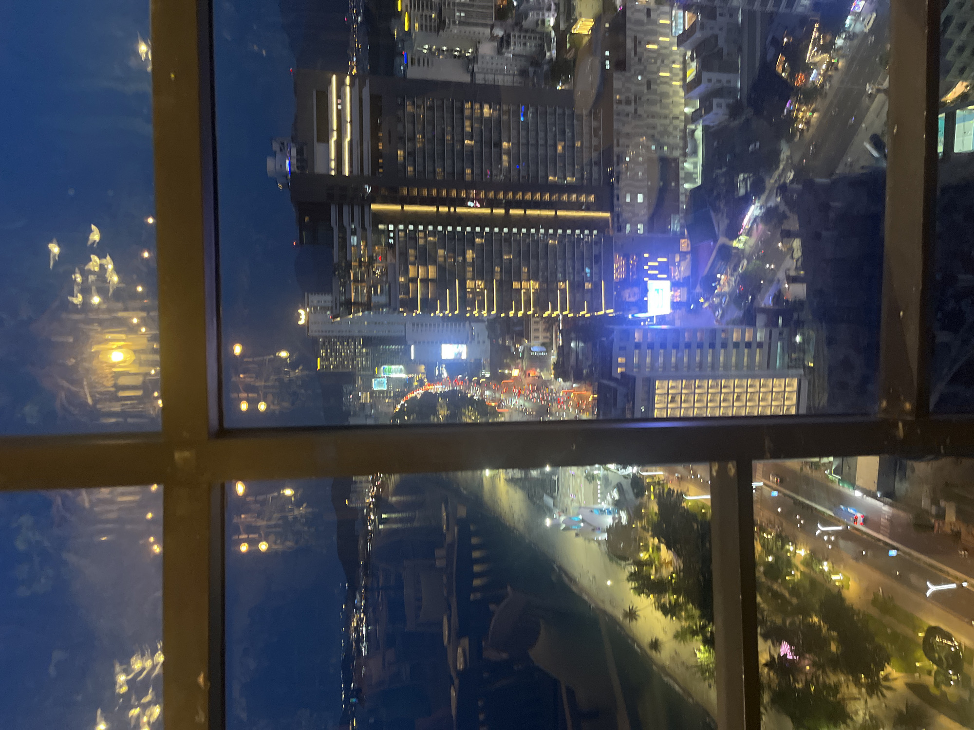 The image shows a nighttime aerial view of the coastal city of Nha Trang, Vietnam, as seen through a window with a dark frame. The city lights illuminate the scene, with buildings, roads, and a coastline visible. The foreground is dominated by the window frame, which is divided into four panes by dark metal bars.
In the upper left pane, reflections of lights are visible on the glass, creating abstract patterns against the dark sky and distant lights. Some of these reflections appear as clusters of small, bright orbs.
The main body of the image, visible through the lower and right panes, displays a bustling urban environment. Tall buildings with illuminated windows are prominent, with one particularly large, modern skyscraper featuring a distinct pattern of golden lights along its facade. A busy road with car headlights and taillights can be seen winding through the city, indicating traffic flow. The coastline is suggested by a darker strip of land bordering the illuminated city, with what appears to be a beach or promenade running alongside it. A few palm trees are visible along this strip, with some illuminated. Further in the distance, more city lights extend towards the horizon, and mountains can be faintly discerned against the night sky. The overall impression is one of a vibrant, well-lit city at night.