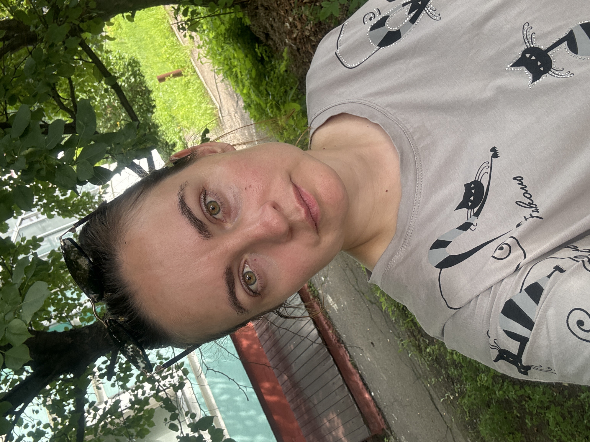 A middle-aged woman with light brown hair tied up and sunglasses perched on top of her head takes a selfie outdoors. She is wearing a light gray t-shirt with a pattern of black cats and the word "Fabulous" written on it. The background shows trees with green leaves, lush green grass, a building with a turquoise and red-brown facade, and a paved path. The scene appears to be a residential area in Blagoveshchensk, Russia, given the architectural style of the building and the surrounding vegetation.