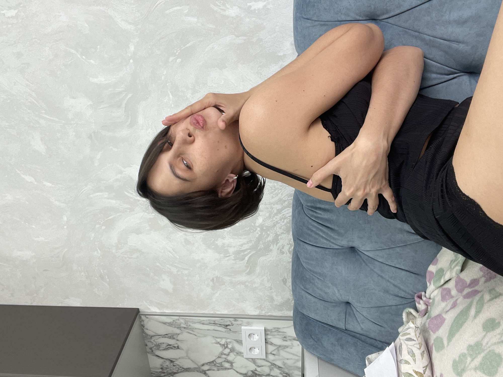 A young woman with short dark hair is reclining on a light blue sofa or armchair, with a white patterned blanket partially visible on the right side of the frame. She is wearing a black sleeveless top and dark shorts. Her right hand is touching her face, and her expression is directed toward the viewer.
The background includes a light gray wall with a marble-like texture. A gray cabinet is visible in the lower left corner. A white double electrical outlet is present on the wall near the floor.
There is no explicit information indicating the location is in Izhevsk, Russia.