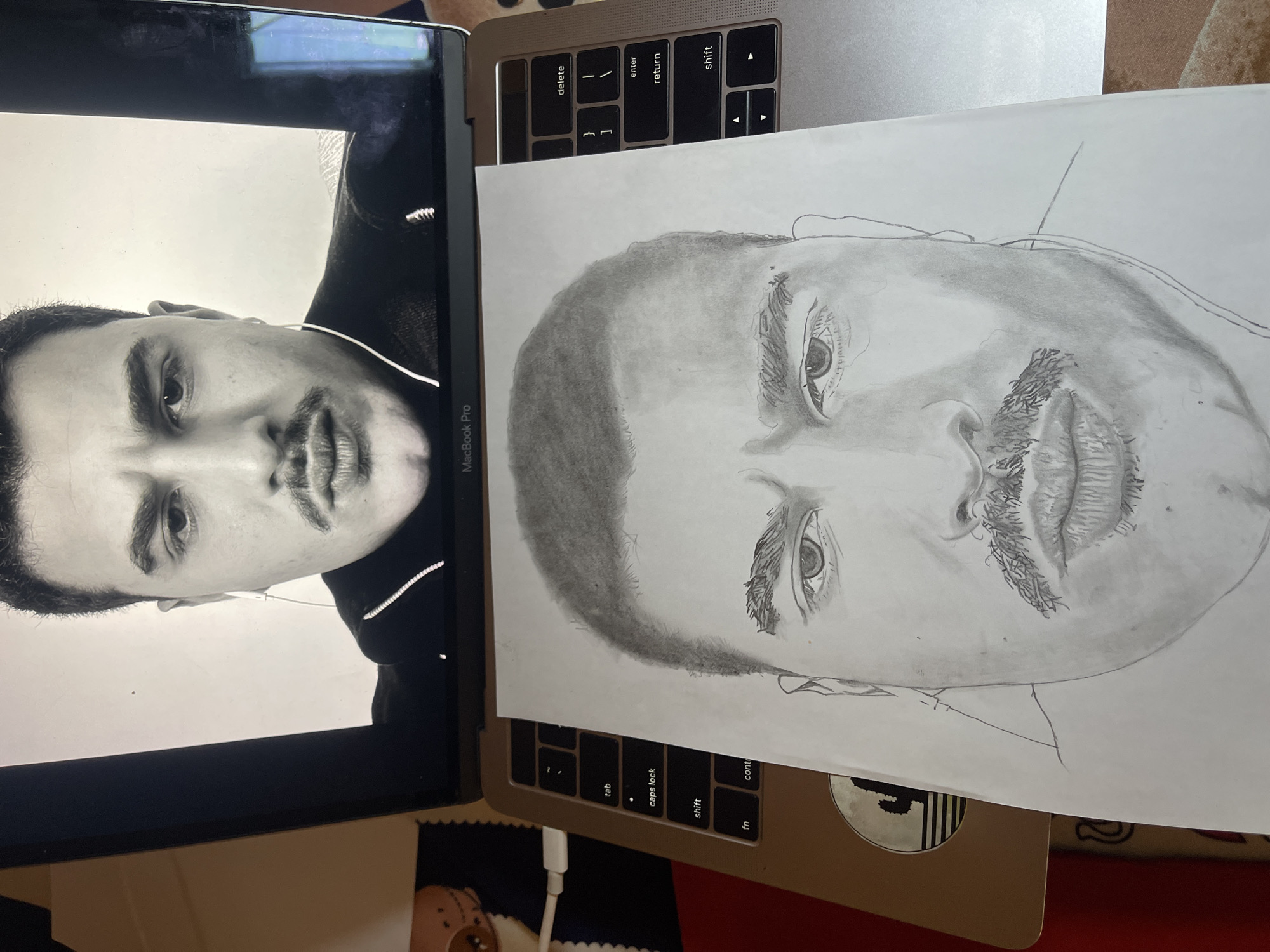 This image depicts an indoor scene in Jos, Nigeria, showing a silver MacBook Pro laptop with an open lid. The laptop's screen displays a black and white head-and-shoulders image of a man with dark hair, prominent eyebrows, a mustache, and a serious expression, wearing a dark top and white wired earphones. A sheet of white paper featuring a pencil portrait of a man's face, also with dark hair, prominent eyebrows, and a mustache, is laid flat on the laptop's keyboard, obscuring most of the keys. A white USB-C charging cable is plugged into the left side of the laptop. A circular sticker with a stylized cactus is visible on the laptop's left palm rest. The laptop and drawing are situated on a surface, with a segment of a multi-patterned fabric visible to the left.
