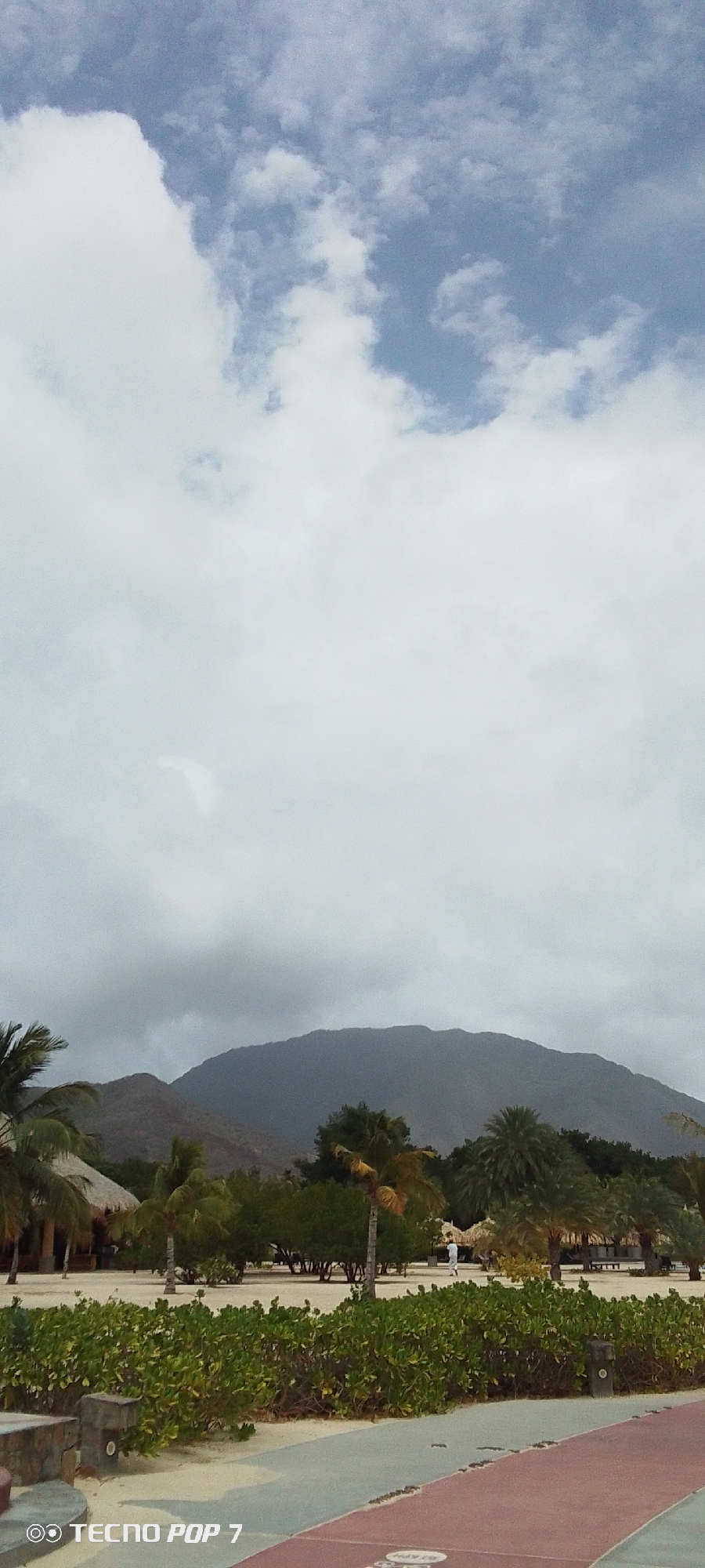 The image shows a view from Pedro González, Venezuela. In the foreground is a partially visible walkway with patches of red and gray. The middle ground consists of low green shrubbery and palm trees interspersed with beach umbrellas. Further back, a mountain range is present, partially obscured by heavy clouds. The sky is predominantly gray with some patches of blue peeking through the clouds. A person is visible walking in the background near the beach umbrellas.