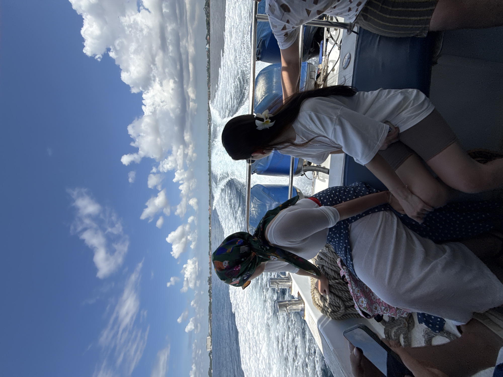 The image shows a group of people on a boat, presumably a tour boat, on the water near the city of Denpasar, Indonesia. Two women are visible near the boat's edge, looking out at the water. One woman has a flower in her hair and wears a white shirt. The other woman wears a patterned headscarf, a white shirt, and a dress. Another person is partially visible wearing a patterned shirt. The boat is moving, as evidenced by the wake in the water. The sky is bright blue with fluffy white clouds. The coastline of Denpasar is visible in the distance. A mobile phone is visible at the bottom of the frame.