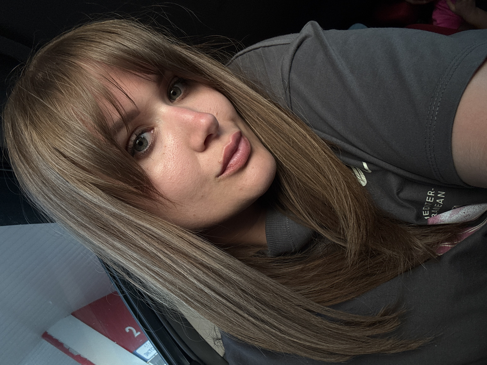 A woman with light brown hair and bangs is posing for a selfie. She is wearing a gray t-shirt with visible lettering partially obscured, potentially reading "MEDITERRANEAN." Her face is angled towards the camera, with a slight tilt of her head. The background is dark and indistinct, suggesting an indoor setting. A corner of a box or package is visible on the left side of the frame. No information from Vinogradny, Russia, is discernible from the image.