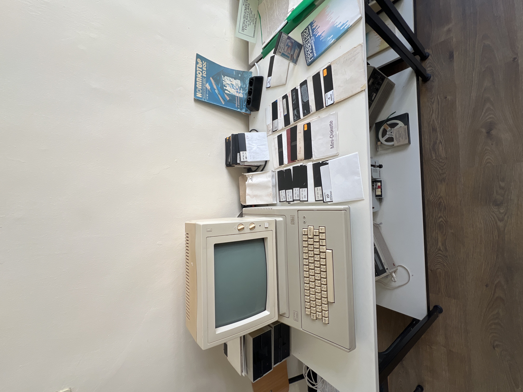 In the city of Pravets, Bulgaria, on a white desk with black legs, sits a vintage Pravets 8D personal computer. The computer features a beige monitor tilted upwards, a beige keyboard with Cyrillic characters, and two floppy disk drives stacked vertically to the left of the monitor. Scattered around the computer are various items, including stacks of floppy disks in protective sleeves, some labeled with text that appears to be in Cyrillic. To the right of the computer, a blue magazine with Cyrillic text and an image of a computer is propped up. Another green booklet or magazine with Cyrillic text is visible further to the right. Beneath the desk, a reel-to-reel tape recorder is partially visible. The background is a plain, light-colored wall. The overall scene depicts a collection of retro computing equipment, possibly in a home or office setting in Pravets.