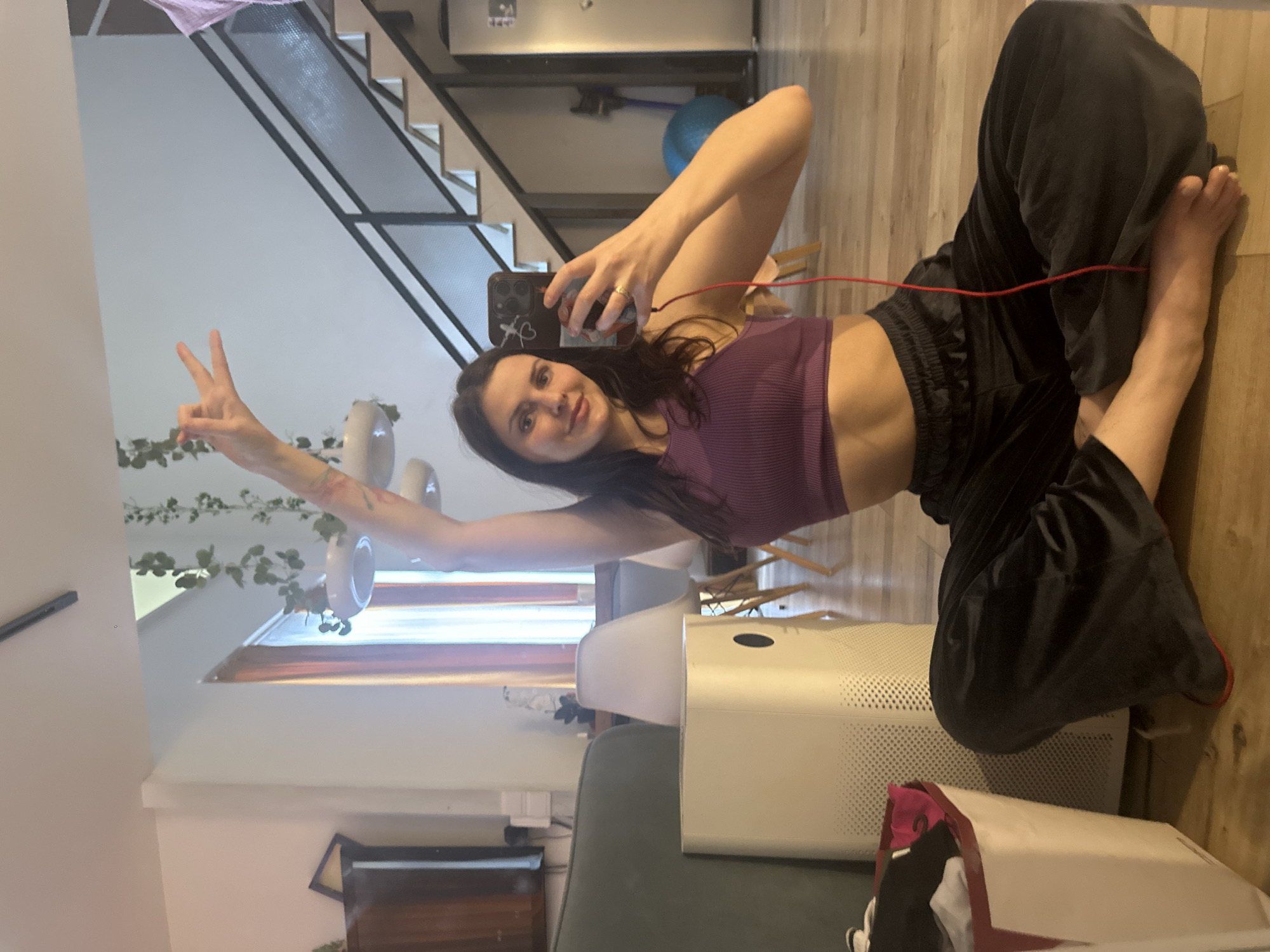 This media file shows a young woman with dark hair taking a selfie in a room. She is wearing a purple crop top and black pants. She is sitting on the floor, with one leg extended and the other bent. She is holding a phone in her right hand and making a peace sign with her left hand. The room appears to be a modern apartment with wooden floors and a staircase in the background. There is also an air purifier and some plants visible in the scene. The lighting suggests it is daytime, and the overall mood is casual and relaxed. No visible text is present in the image.