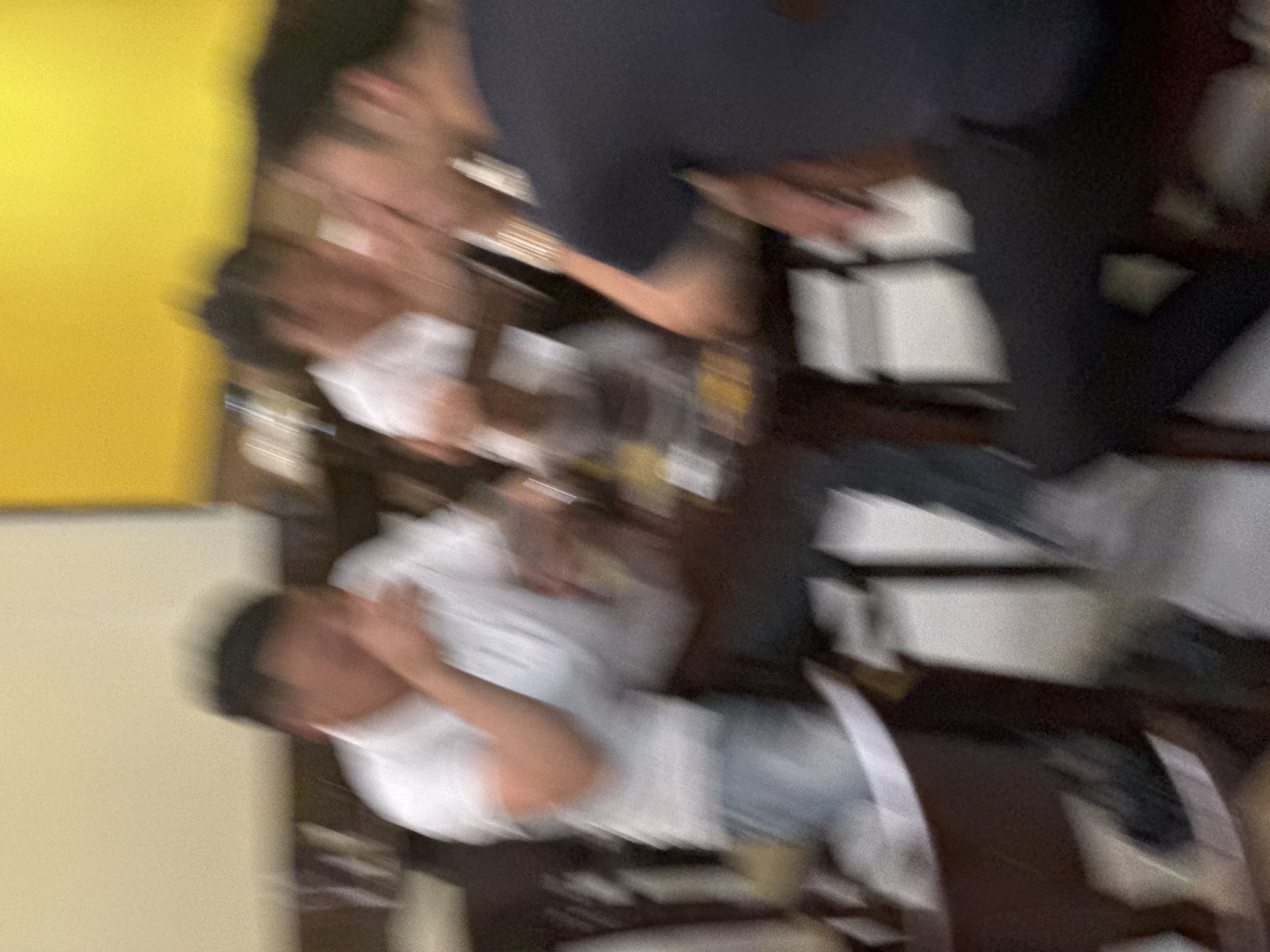 The image, captured from a slightly elevated angle, depicts a group of individuals seated around a table in what appears to be a casual dining setting. The scene is characterized by significant motion blur, making precise identification of features challenging.
In the foreground, a person in a dark t-shirt is visible, their form blurred as they lean forward. To their left, another individual, also with blurred features, is wearing a white t-shirt and light-colored pants. Their arm is extended, and they appear to be interacting with someone across the table.
Further into the background, more blurred figures are discernible. One individual in a white shirt is leaning on their hand, their face obscured by motion. Another person, partially visible, is in the process of leaning over the table.
The environment suggests an indoor location, possibly a restaurant or a similar establishment. The background features a bright yellow wall or ceiling, contrasting with the darker tones of the furniture and the people. The table itself is laden with what appear to be glasses and possibly some food items, though details are indistinct due to the blur. The overall impression is that of a lively gathering, possibly a meal or drinks, with a sense of movement and interaction.
Based on the provided context, the location is identified as Hoa Lư Ward, Vietnam.