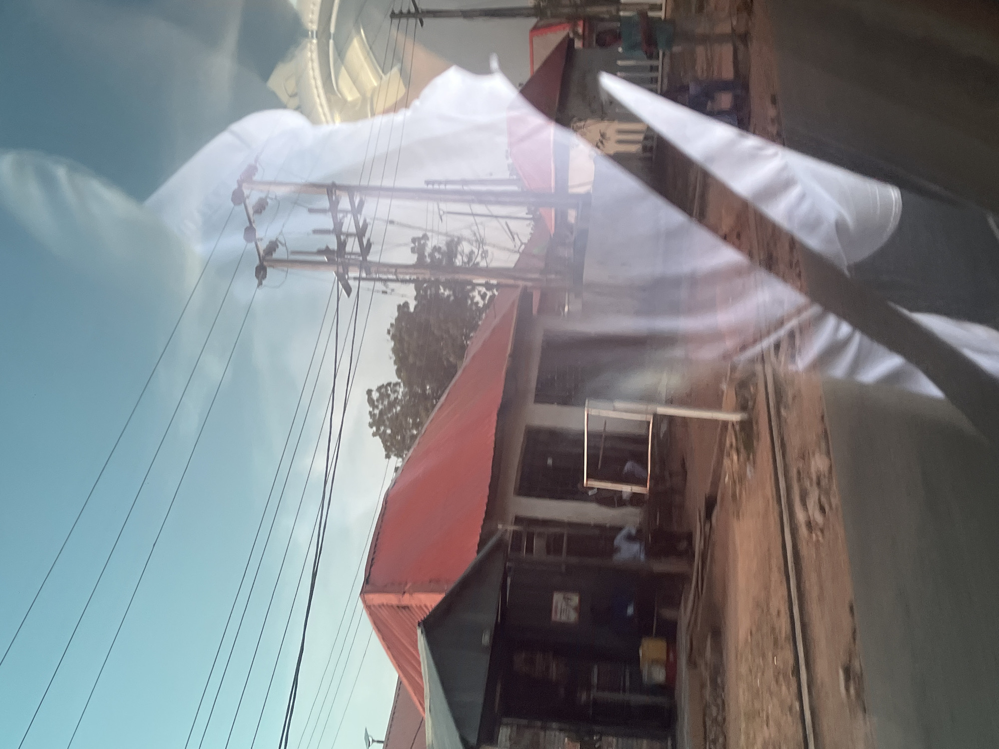 The image shows a street scene in Jalingo, Nigeria, with a building featuring a red roof and a dark facade. There are several people visible, though they are mostly indistinct due to the lighting and reflections. Some are standing outside the building, while others appear to be inside. Electrical wires are strung across the sky, suggesting an urban or semi-urban environment. The sky is a pale blue, indicating it might be daytime, though the reflections on a surface, possibly a window or vehicle, obscure some details and create a layered effect. The overall impression is of a busy, everyday scene with elements of both residential and commercial activity. The reflections also show a large white, possibly fabric or plastic, object, which is difficult to identify precisely.