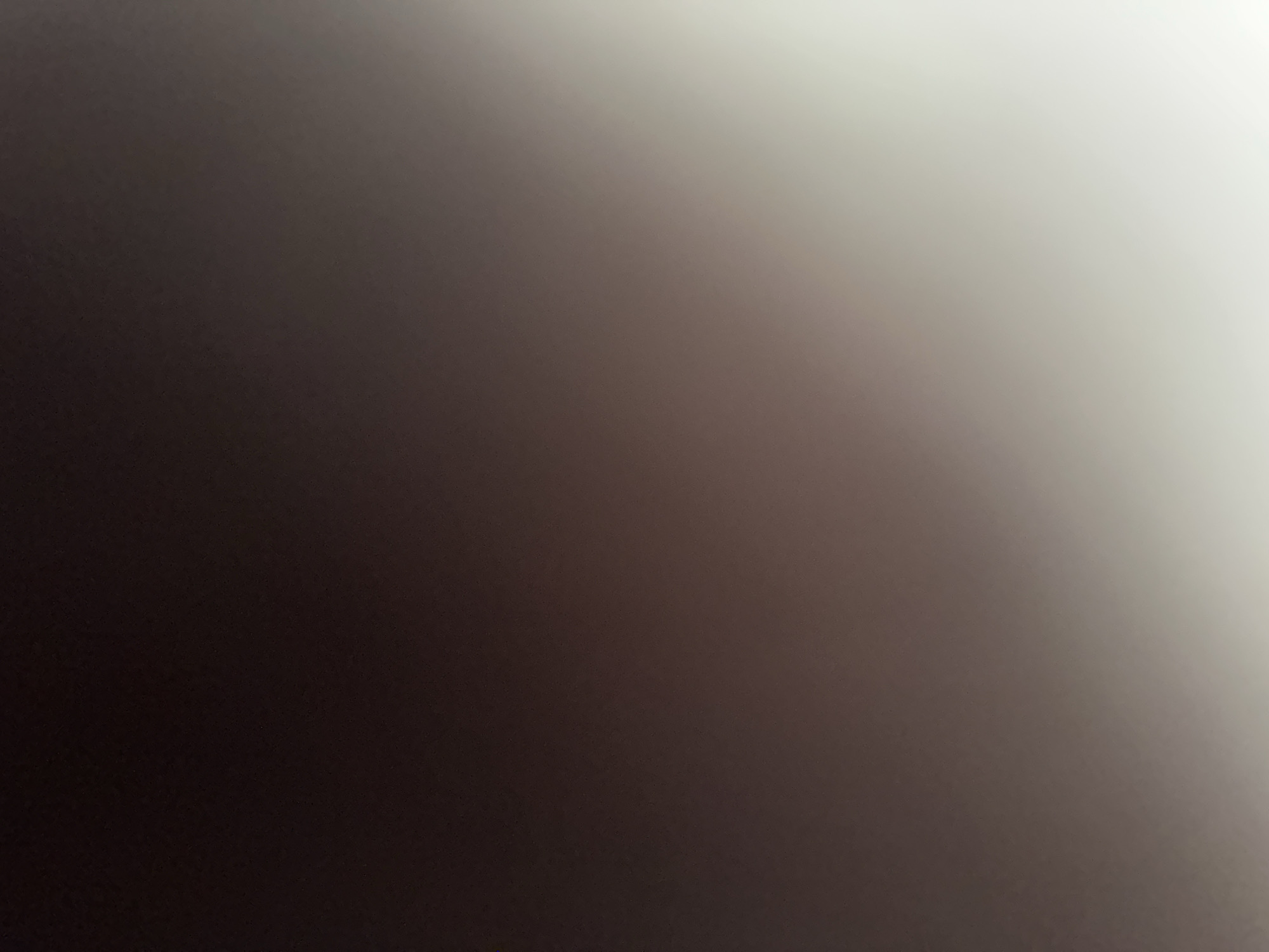 This is a photograph with a dark gradient effect. The left side of the image is a deep, rich brown, which gradually lightens towards the right. The right side of the image transitions into a soft, light grey or off-white. There are no discernible subjects, objects, scene, or setting. No activity or event is taking place. The image is abstract due to the lack of specific visual elements. There are no notable details such as time of day, weather, location cues, or emotions. No text is visible in the image. The photograph appears to be a close-up of a textured surface or a deliberate lighting effect, creating a smooth, abstract gradient.