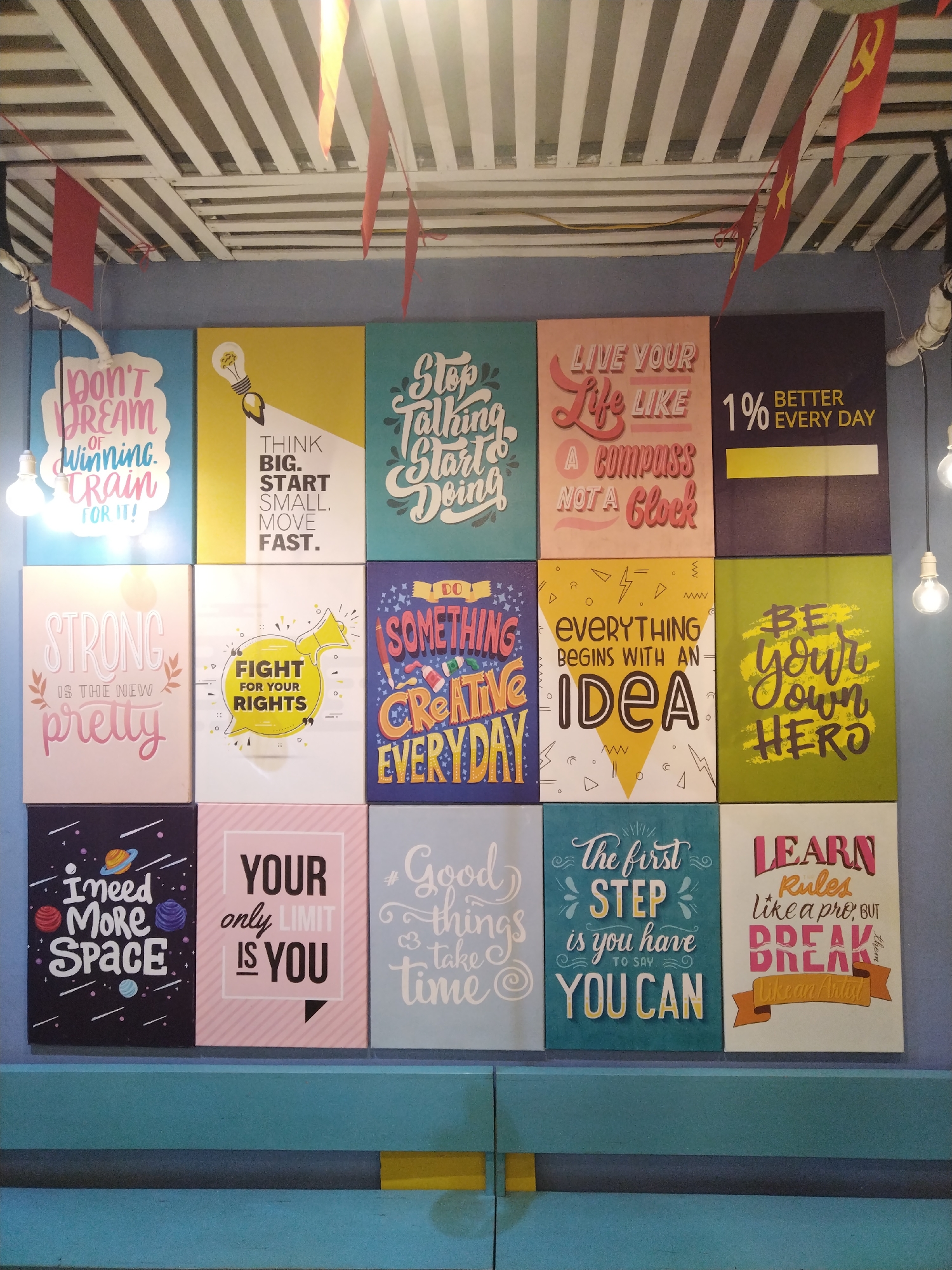 The image shows a wall decorated with 12 motivational posters in a 3x4 grid format. The posters feature various inspiring quotes and illustrations in diverse color schemes. Red and yellow flags hang from the ceiling above the posters, suggesting a possible theme or location. Light bulbs are also visible, providing illumination. A horizontal beam with blue and yellow elements runs along the bottom of the image. There is no evidence of this being Bai Chay Ward, Vietnam.