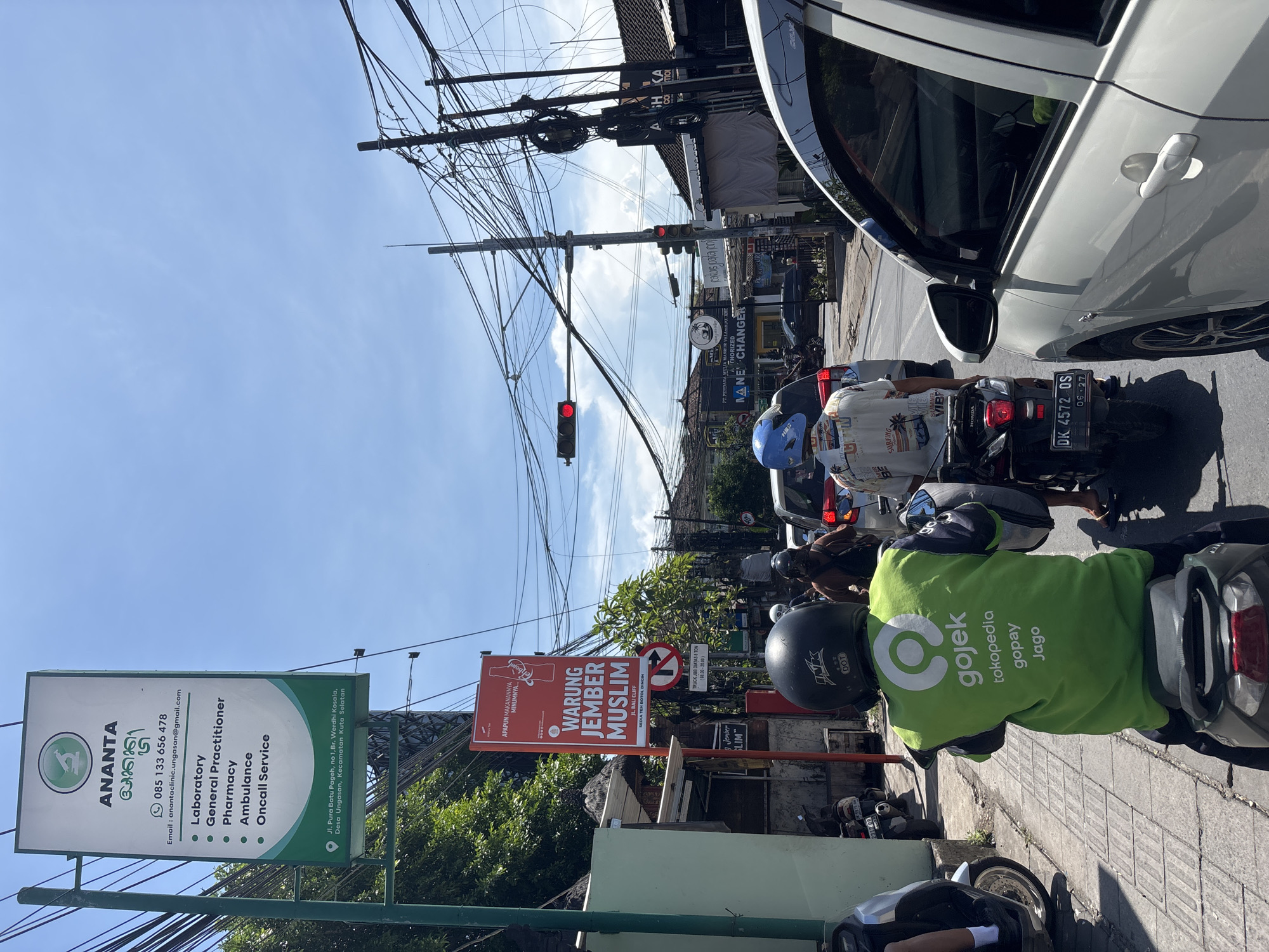 The image captures a street scene in Kuta Selatan, Indonesia. A Gojek driver in a green jacket is waiting on a motorbike near the curb. Beside the driver are two street signs, one for "ANANTA," a clinic, and another sign that says "WARUNG JEMBER MUSLIM." The traffic light is red. Another motorbike waits behind the Gojek driver. A white car is partially visible on the right. Overhead, there's a complex web of tangled power lines against a blue sky. Buildings and various commercial signs line the street, including a "MONEY CHANGER" sign.