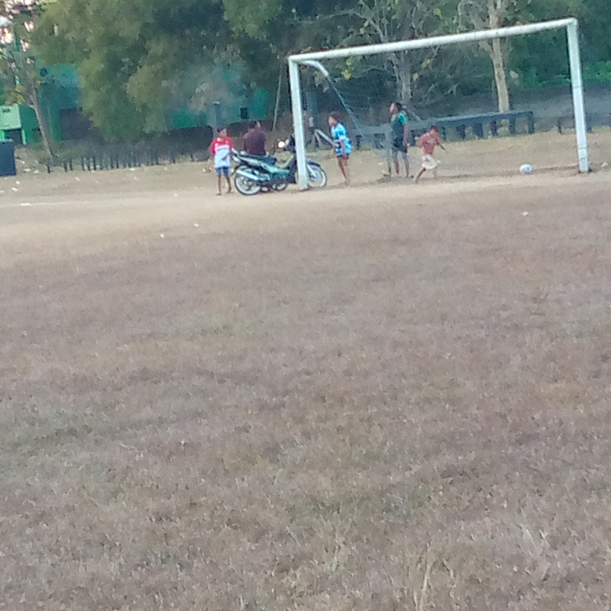 The image depicts a scene in Pacing, Indonesia, likely an open field with a soccer goal. Several individuals are present. To the left, a young person in a red and white shirt and blue shorts stands near a fence and some trees. A motorcycle is parked to the right of this individual, with at least one person seated on it wearing a brown shirt. Further to the right, two individuals are near the soccer goal, one in a blue and white patterned shirt and another in a dark shirt. To the right of the goal, another individual in a red shirt is in motion, appearing to be kicking a soccer ball. A soccer ball is visible on the ground near this person. The ground is dry and covered with sparse vegetation. Trees and some structures are visible in the background.