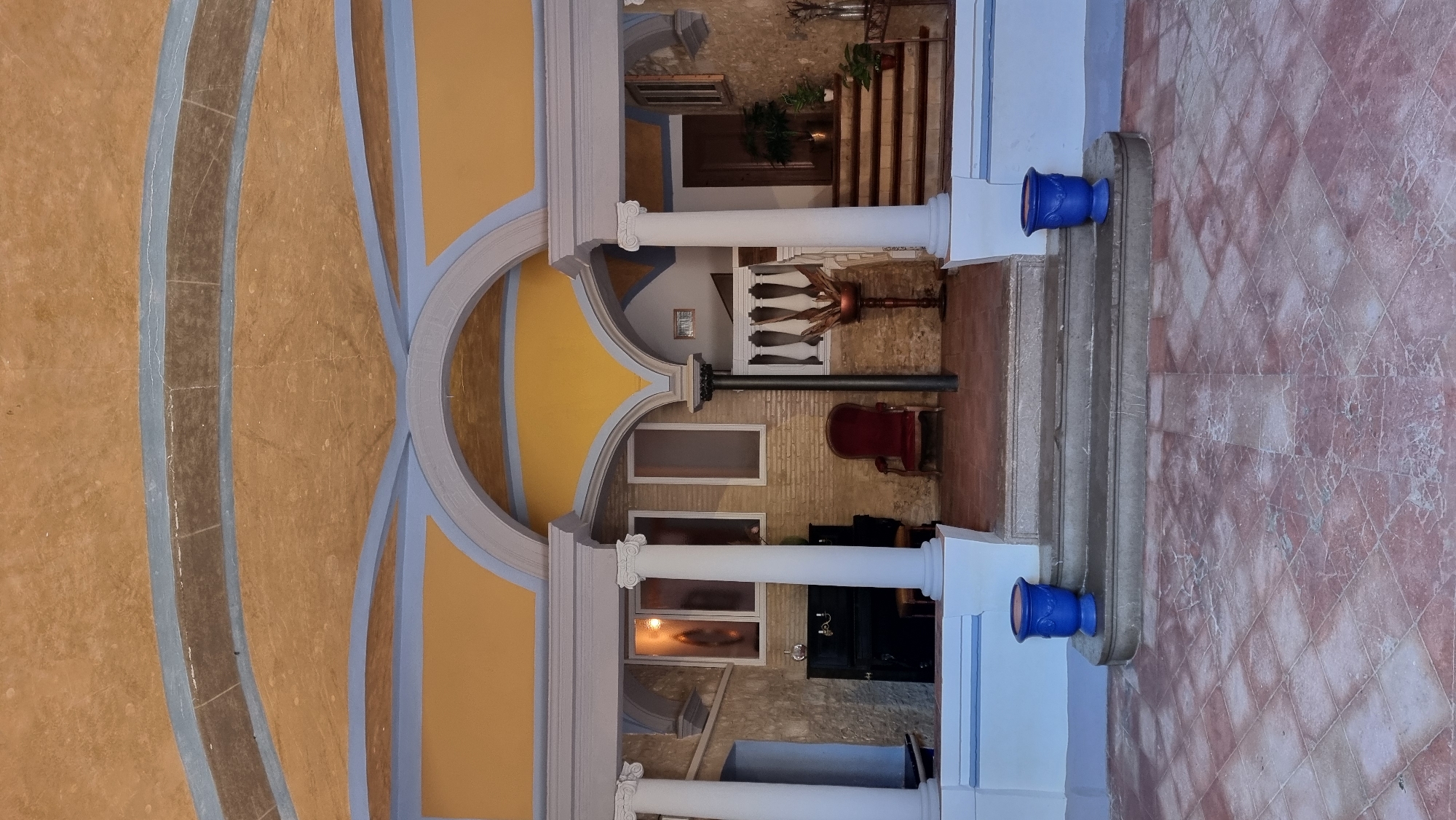 The image depicts an outdoor covered area in front of a building, likely in Castelló d'Empúries, Spain. The architectural style features a yellow and blue arched ceiling with decorative molding. The space is supported by white classical columns. In the foreground, there are stone steps leading up to the covered area, with two blue planters on either side of the bottom steps. The ground is paved with reddish-brown square tiles.
Inside the covered area, to the left, is a black upright piano positioned against a stone wall. Beside the piano are tall, narrow windows with frosted glass, one of which shows a faint reflection of a figure. A single red velvet armchair is visible in the center. To the right, a white balustrade lines a staircase leading upwards. On the steps, there are potted plants and a decorative wooden stand holding ornamental items. Further to the right, a dark wooden door is visible. The walls of the building appear to be made of textured stone or brick. The overall impression is of an entryway to a residence or historical building.