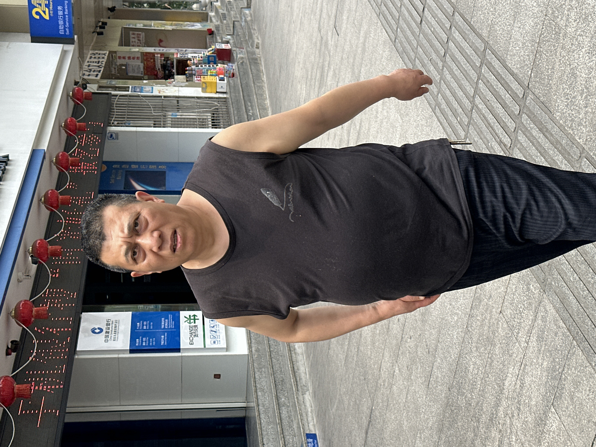 The image shows an Asian man, wearing a black tank top and black trousers, standing on a sidewalk in Jiulongpo District, China. Behind him is a branch of the China Construction Bank, adorned with red lantern-like lights and bank signage in Chinese. There are also advertisements in Chinese. The ground appears to be paved with gray stone.