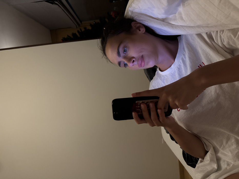 A fair-skinned young woman with dark hair looks at a smartphone held horizontally in her hands. Her face is illuminated by the device's light, suggesting a low-light environment. She's wearing a white t-shirt, and a white pillow or bedding is positioned behind her head. The background is a plain wall, partially illuminated. The overall impression is one of quiet, casual screen time.