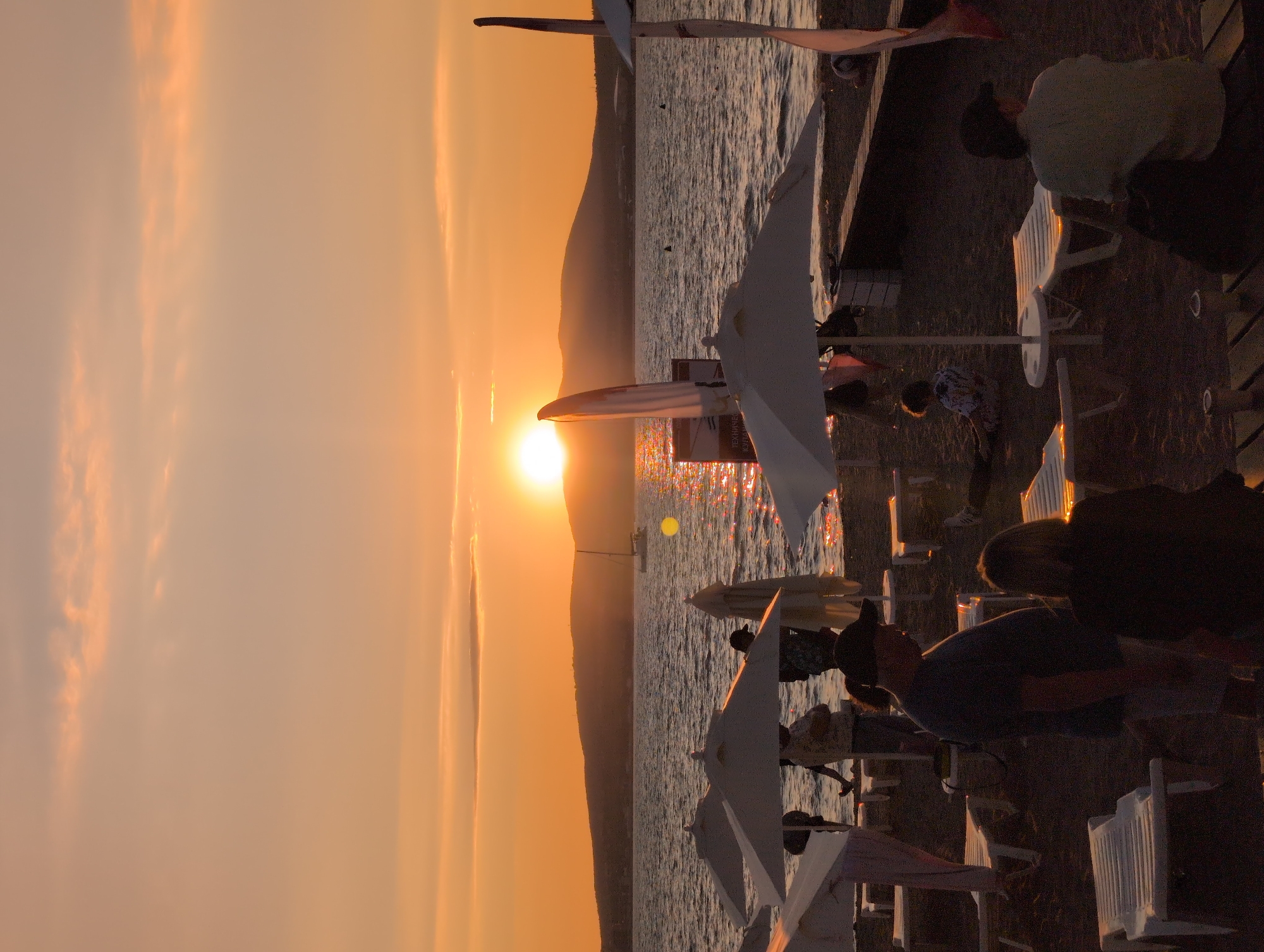 The image depicts a sunset scene on a beach in Gelendzhik, Russia. The sun is setting behind a chain of hills that form the coastline, casting a warm orange glow across the sky and reflecting on the rippling water. Several people are visible on the sandy beach, some of whom appear to be relaxing under beach umbrellas, while others are standing or walking. Beach chairs are arranged on the sand, indicating a popular recreational area. A sailboat is visible in the distance on the water. The overall atmosphere is one of a peaceful evening by the sea.