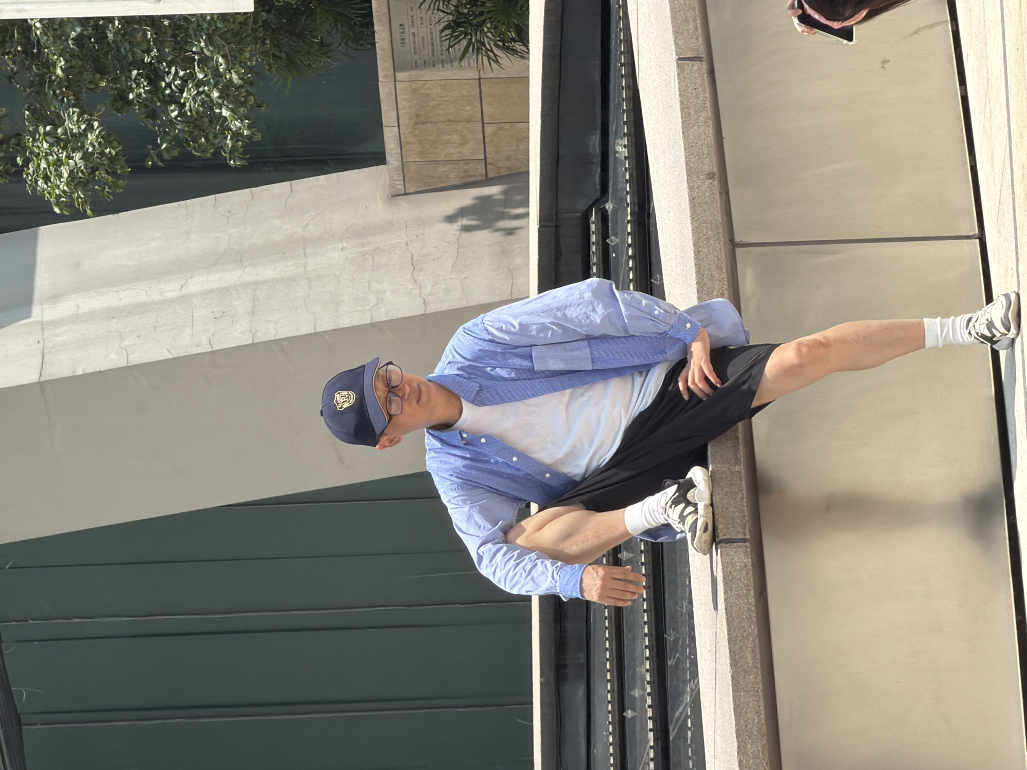 The image shows an East Asian male sitting on a concrete ledge in an outdoor urban environment. He is wearing a blue collared shirt, a white t-shirt, black shorts, a navy blue baseball cap, glasses, white socks, and athletic shoes. His left leg is bent at the knee, with his foot resting on the ledge. His right leg is extended downwards. He is looking to his right and appears to be relaxed.
Behind him, there is a large concrete structure with a curved design, and a dark green vertical panel. To the right of the concrete structure, there is a tree with green foliage and a wall made of stone tiles. In the foreground, there is a water feature or fountain with dark water and small lights. The ground in front of the ledge is paved. The overall setting suggests a public space, possibly a park or plaza within the city of Yuzhong District, China.