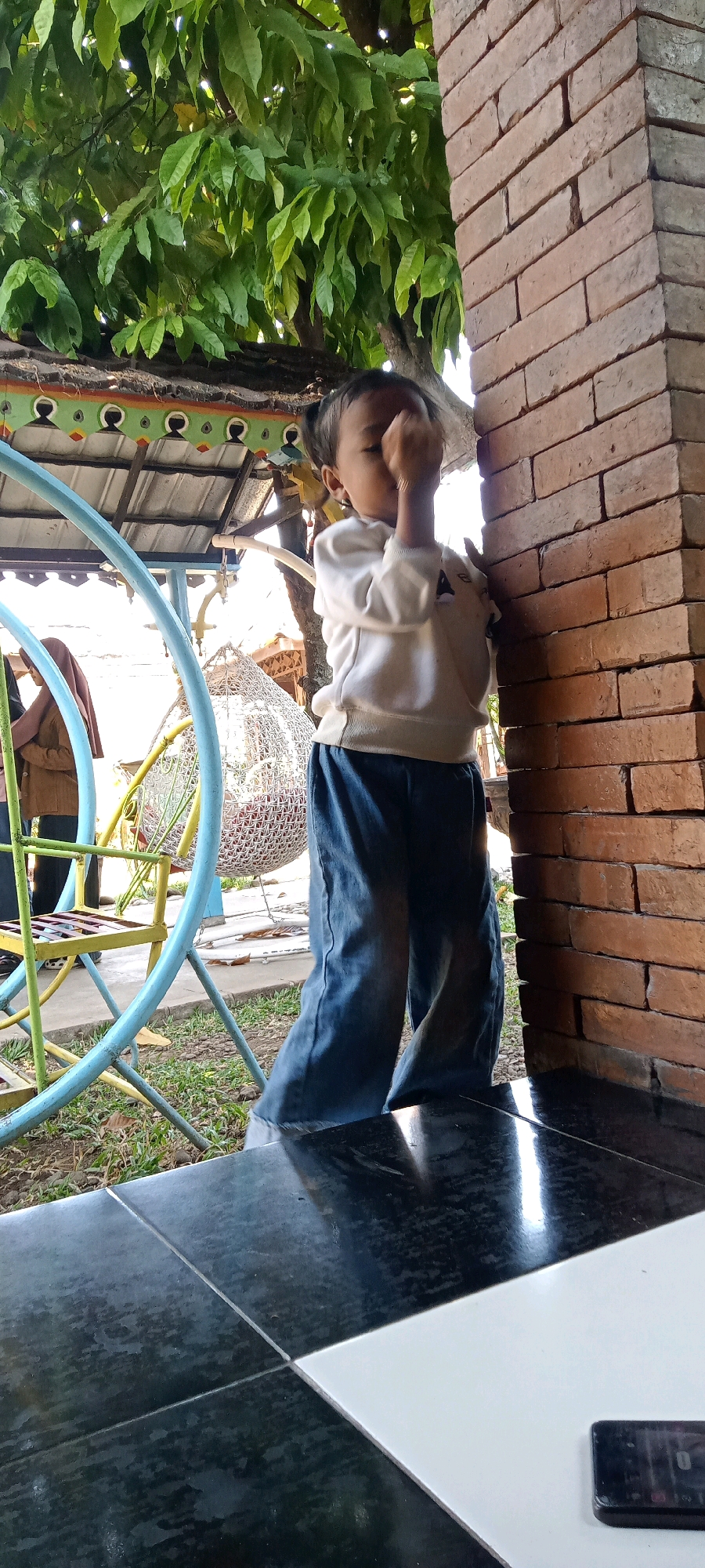 In Senggrong, Indonesia, a young child stands partially obscured by a brick pillar, facing a leafy tree. The child, wearing a light-colored sweatshirt and blue pants, has their hand raised to their face, possibly rubbing their eyes. To the left of the child, a colorful playground structure with blue and yellow accents is visible. A white, spherical hanging chair is also present behind the child, and two other individuals are partially visible in the background. The ground is covered with grass and some fallen leaves. The scene appears to be outdoors, likely in a public space or a backyard.