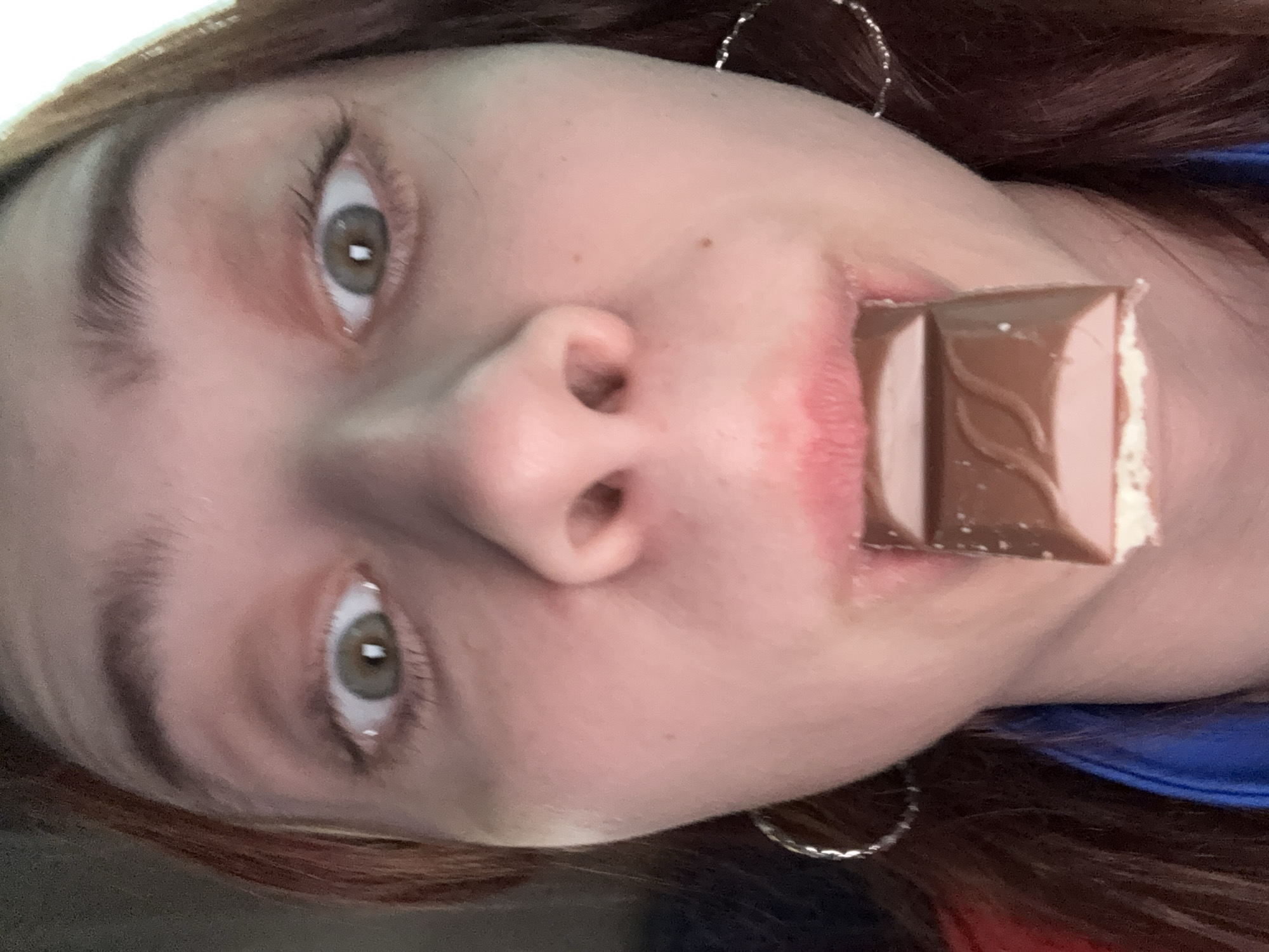 A close-up portrait captures a fair-skinned person with reddish-brown hair and hazel eyes holding a partially eaten chocolate bar in their mouth. The person is wearing silver hoop earrings. The background is out of focus and provides no definitive information regarding location, making it impossible to place the scene in the city of Zvyozdny, Russia.