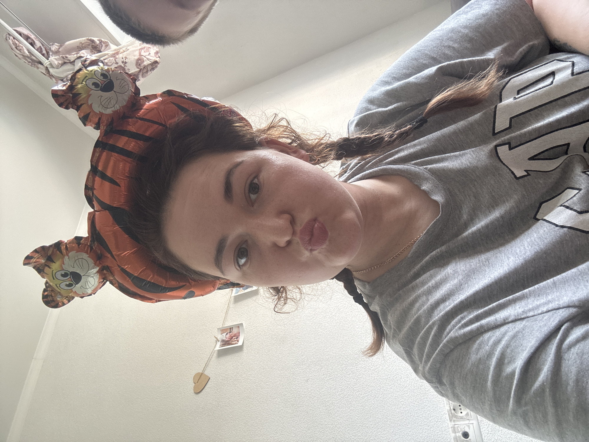 A young woman wearing a grey t-shirt with "GAP" printed on it, and a tiger-shaped balloon headband, poses for a selfie against a white wall. She has her hair in braids and is puckering her lips. Two small photos are clipped to a string on the wall behind her. An electrical outlet is visible on the lower left of the image. The scene appears to be indoors, likely in a residential setting, and there is no indication of Chelyabinsk, Russia.